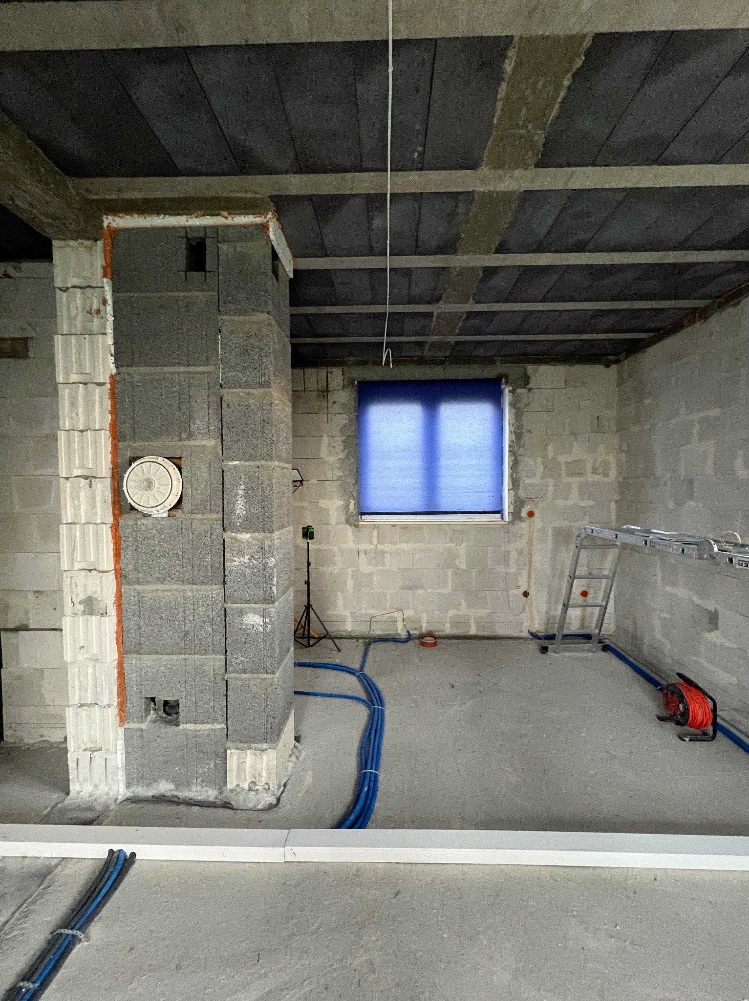 Surowy stan wnętrza w trakcie prac. Widoczne niebieskie rury instalacji elektrycznej i wentylacja w ścianie z szarego betonu. Laser krzyżowy na statywie.