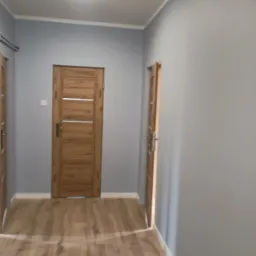 Wnętrze korytarza po remoncie: drewnopodobna podłoga, szare ściany, dwoje drzwi wewnętrznych z poziomymi, srebrnymi akcentami i biała listwa przypodłogowa.