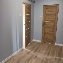 JD Usługi Remontowo Budowlane Jerzy Delekta - Jasne panele podłogowe w wyremontowanym pokoju z szarymi ścianami i dwojgiem drzwi z drewnopodobnymi okleinami, jedno skrzydło otwarte.