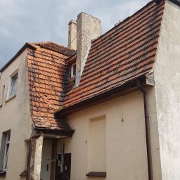 Roof 66 Łukasz Trzpiot - Dwupiętrowy budynek z czerwonym dachem z dachówki ceramicznej, widoczne uszkodzenia i ślady po naprawach na elewacji, dwa kominy z widocznymi ubytkami tynku, rynna w kolorze brązowym, schody...