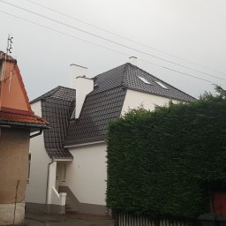 Roof 66 Łukasz Trzpiot - Dom z nowym, ciemnym pokryciem dachowym z dachówki ceramicznej, widoczne dwa okna dachowe i dwa białe kominy, sąsiadujący z budynkiem z czerwoną dachówką i balkonem, na pierwszym planie gęsty...