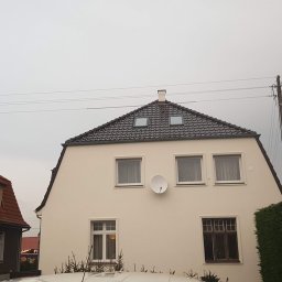 Roof 66 Łukasz Trzpiot - Dwupiętrowy dom z ciemną dachówką, dwoma oknami dachowymi i anteną satelitarną na elewacji, widok z dołu w pochmurny dzień.