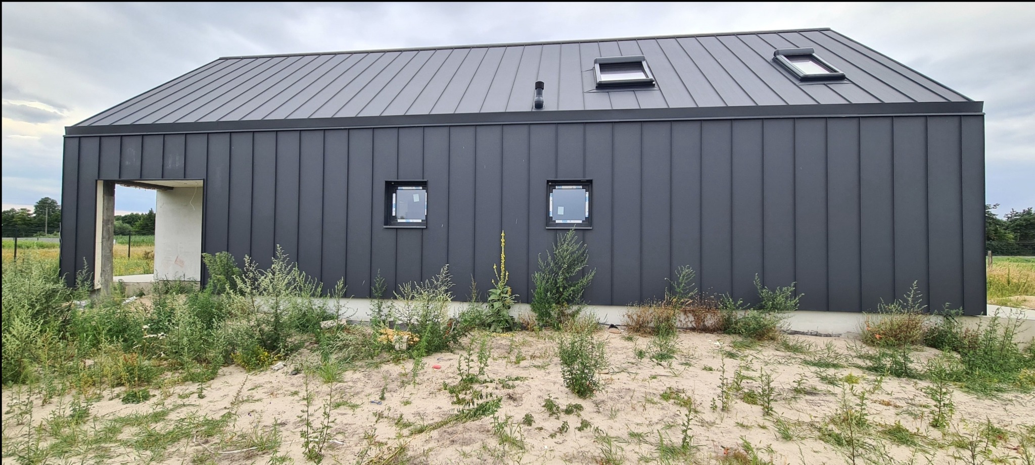 Nowoczesny dom w trakcie budowy z ciemnoszarą blachą na elewacji i dachu, widoczne okna dachowe i dwa małe okna na ścianie, teren wokół budynku zarośnięty chwastami.