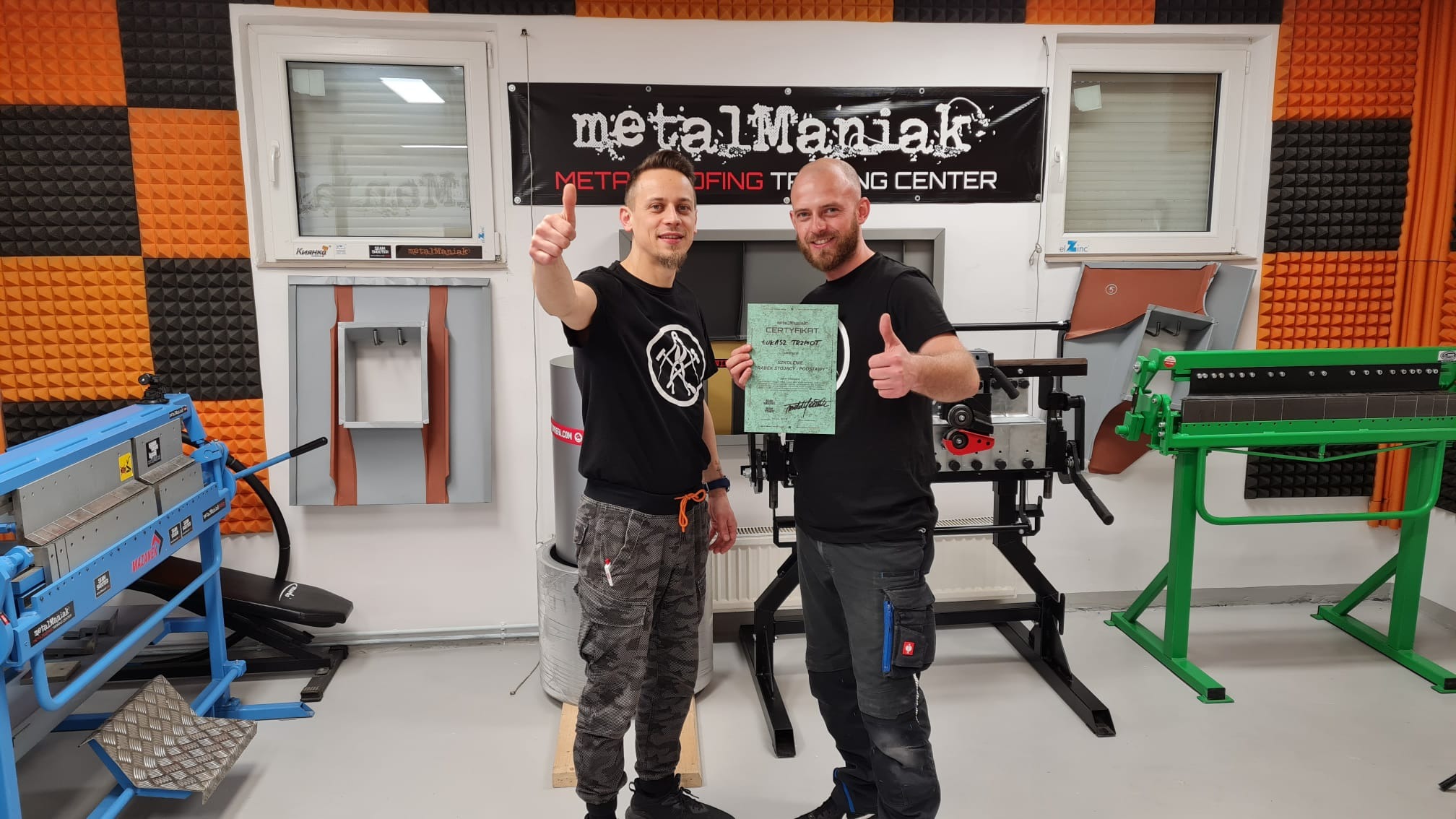 Dwóch mężczyzn z podniesionymi kciukami trzyma certyfikat na tle warsztatu dekarskiego MetalManiak, z maszynami do obróbki blachy i panelem demonstracyjnym na ścianie.