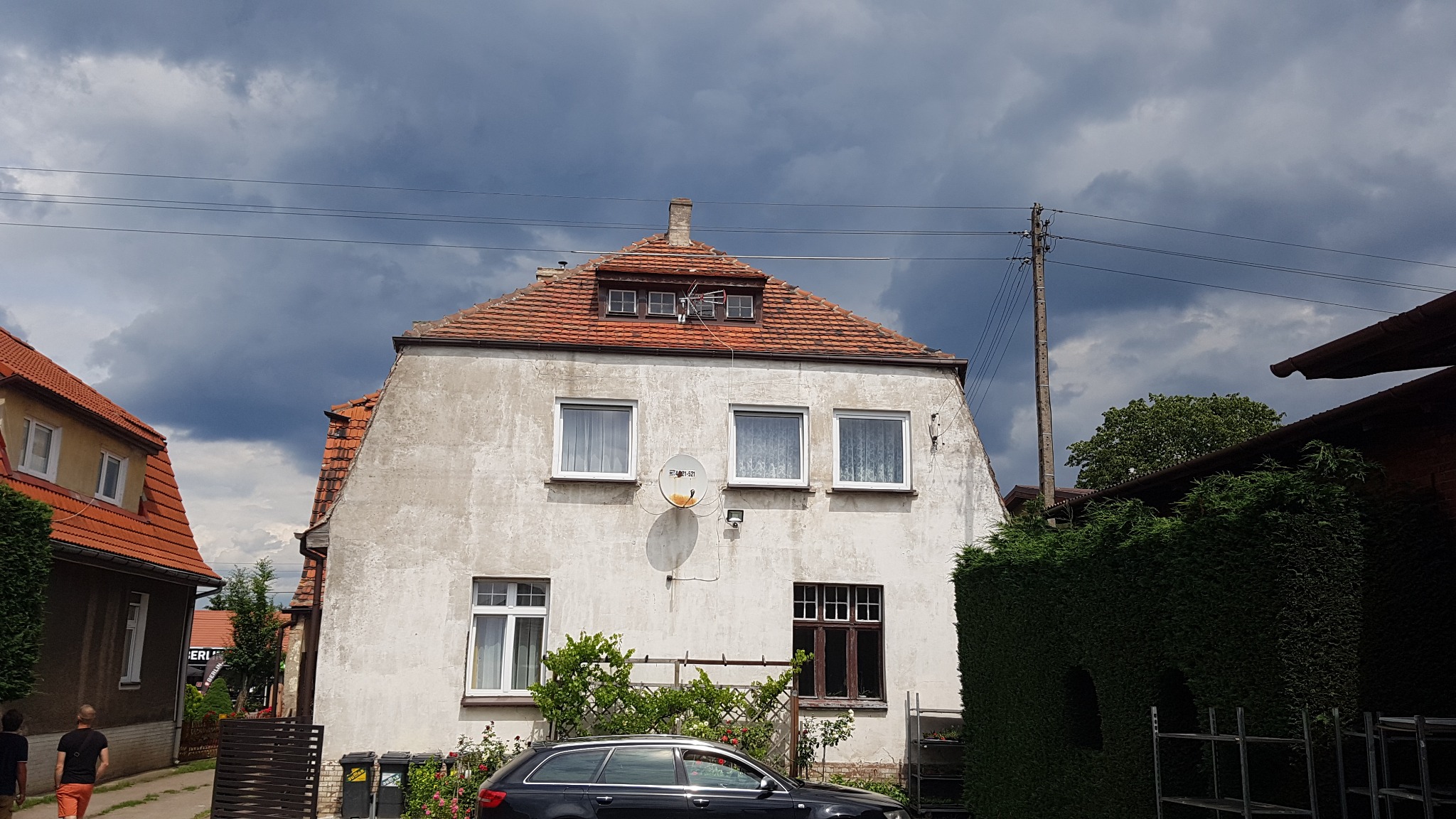 Dwukondygnacyjny dom z pomarańczową dachówką, anteną satelitarną na elewacji i ciemnymi chmurami na niebie.