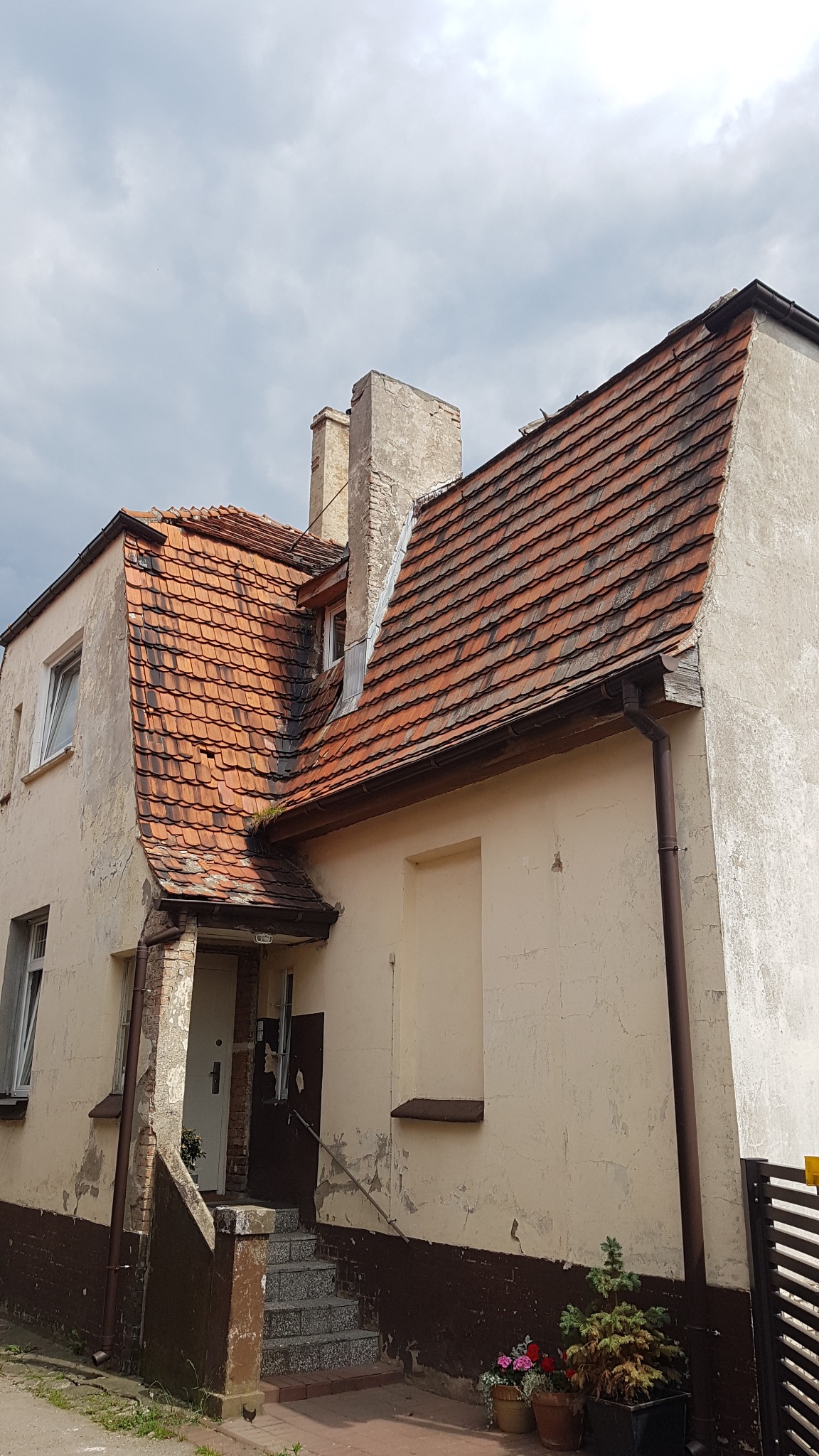 Dwupiętrowy budynek z czerwonym dachem z dachówki ceramicznej, widoczne uszkodzenia i ślady po naprawach na elewacji, dwa kominy z widocznymi ubytkami tynku, rynna w kolorze brązowym, schody...