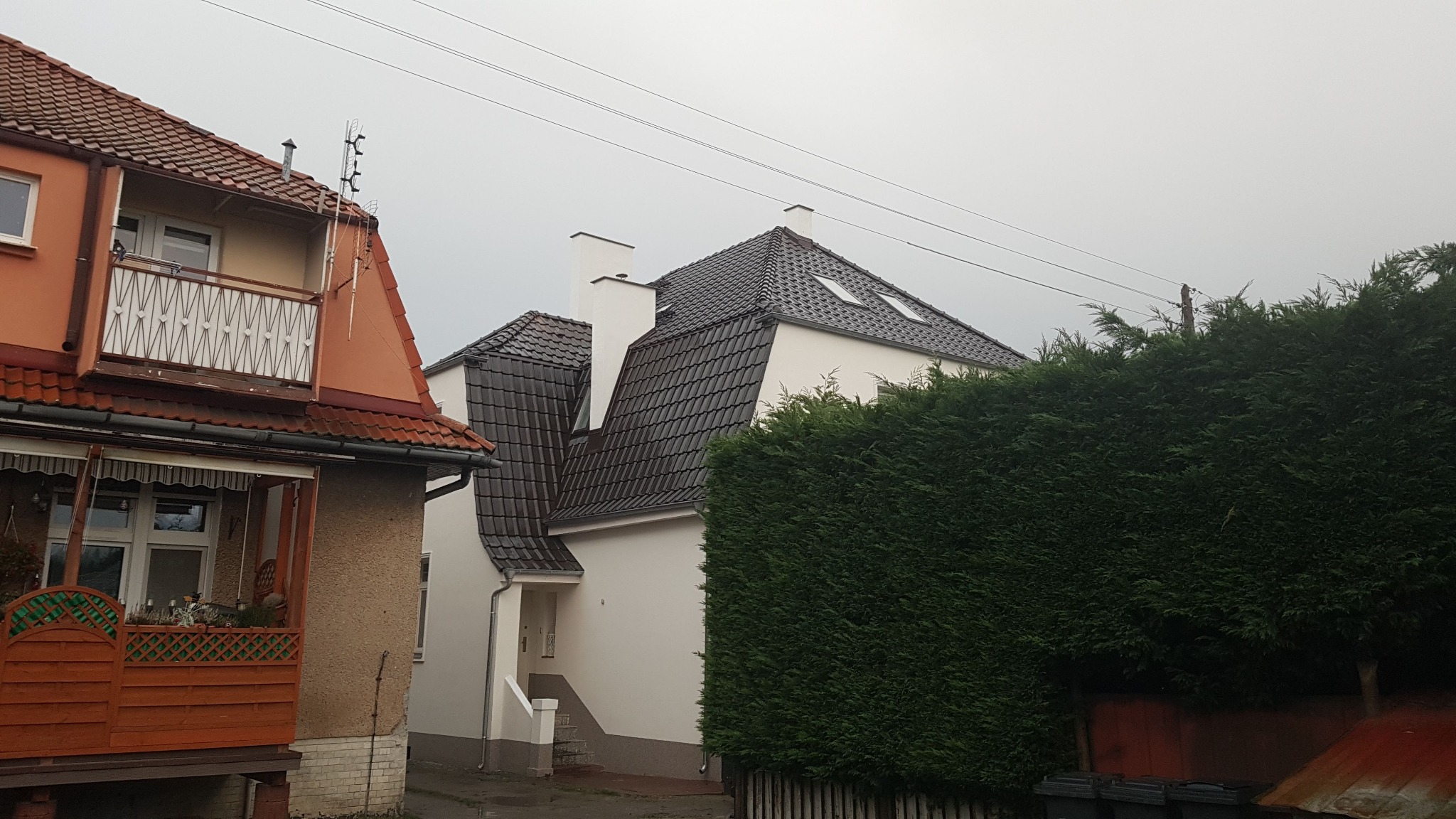 Dom z nowym, ciemnym pokryciem dachowym z dachówki ceramicznej, widoczne dwa okna dachowe i dwa białe kominy, sąsiadujący z budynkiem z czerwoną dachówką i balkonem, na pierwszym planie gęsty...