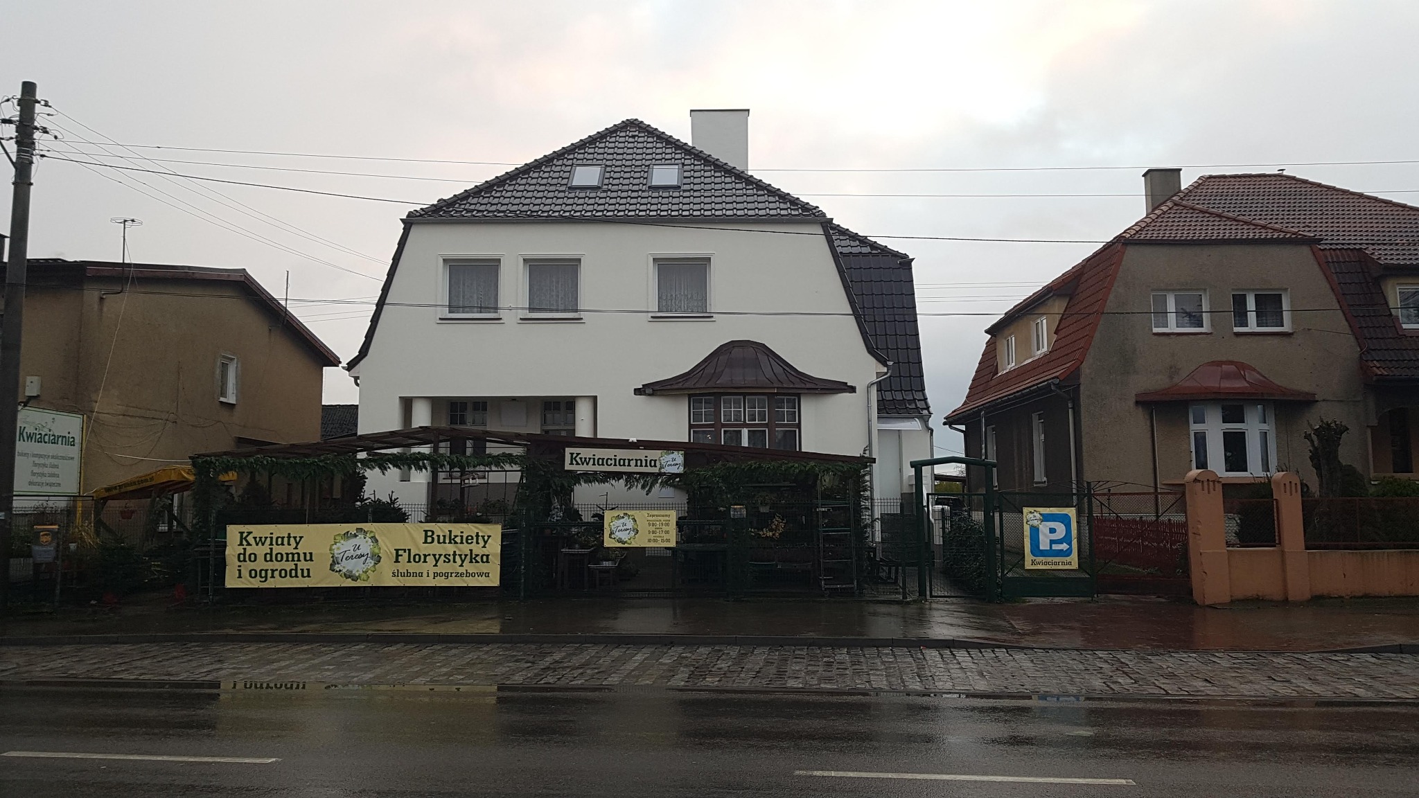 Budynek kwiaciarni z ciemną dachówką i lukarnami, widok z poziomu ulicy w pochmurny dzień.