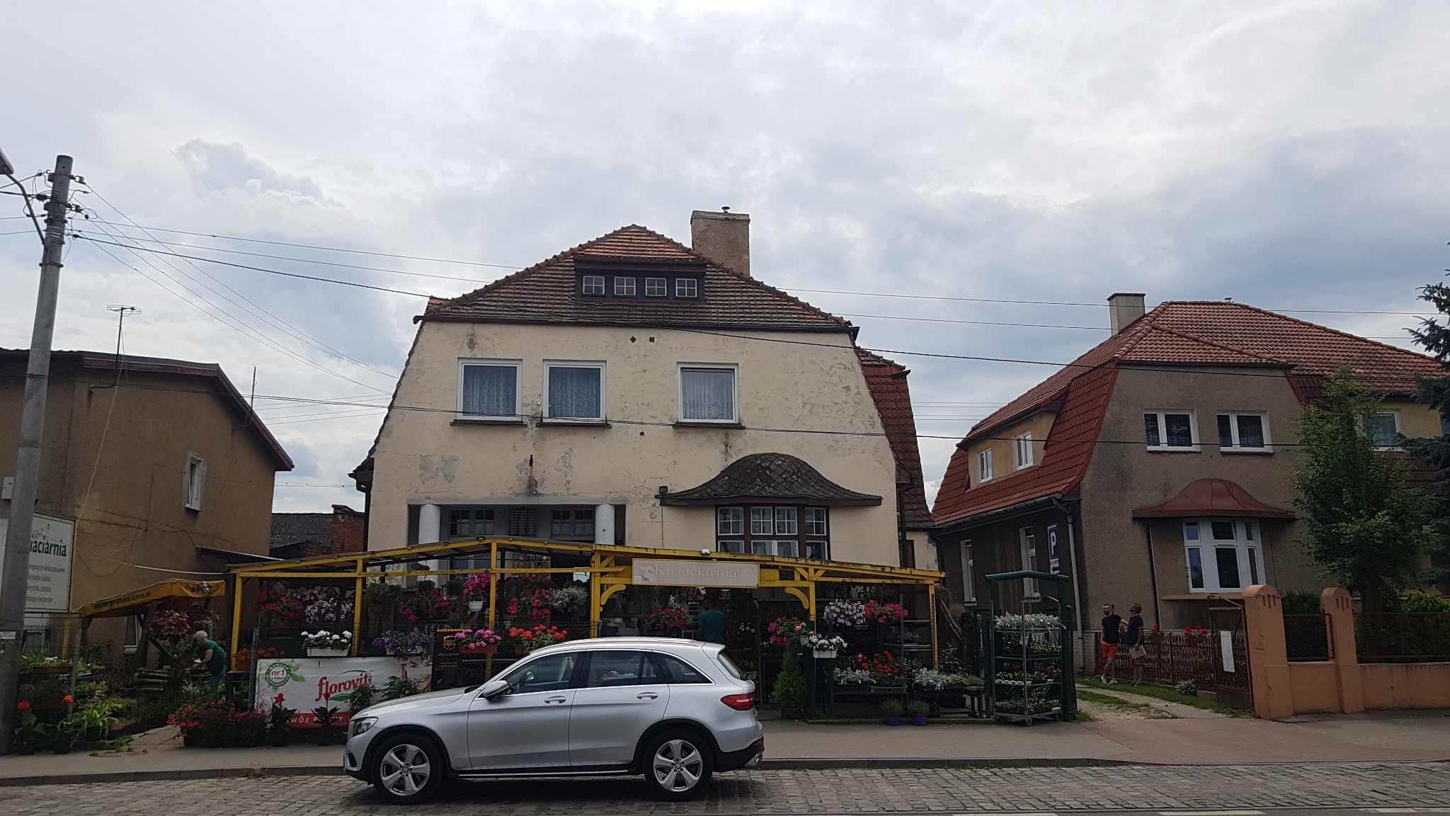 Dwa budynki mieszkalne z czerwonymi dachami, widoczne z ulicy wyłożonej kostką brukową. Przy jednym z budynków, pod żółtą konstrukcją, znajduje się kwiaciarnia z licznymi kwiatami. Na pierwszym...