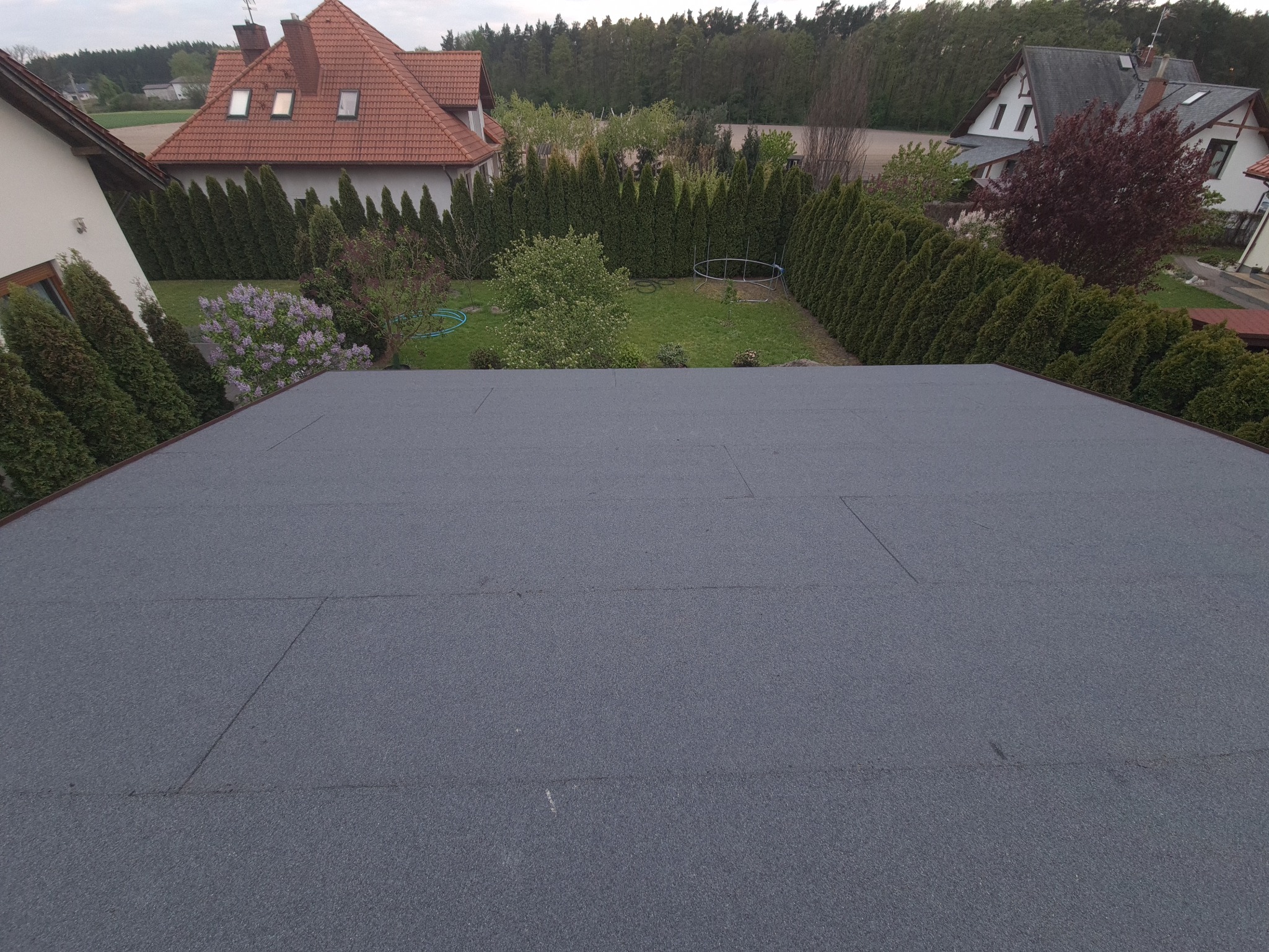 Widok z góry na płaski dach pokryty papą termozgrzewalną, w tle zadbany ogród z tujami, drzewami i domami.