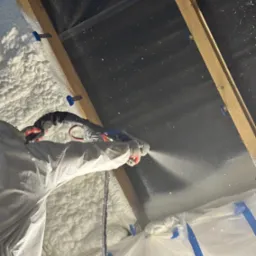 Aplikacja pianki izolacyjnej PUR na poddaszu, widoczne elementy konstrukcji dachu i zabezpieczenia folią. Praca w toku, piana pokrywa powierzchnię.