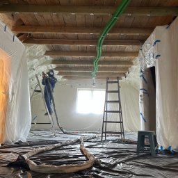 WELSUN ENERGY SPÓŁKA Z OGRANICZONĄ ODPOWIEDZIALNOŚCIĄ - Prace izolacyjne poddasza: aplikacja piany natryskowej przez pracownika w kombinezonie, widoczne zabezpieczenia folią, drewniana konstrukcja dachu i zielone węże doprowadzające materiał.