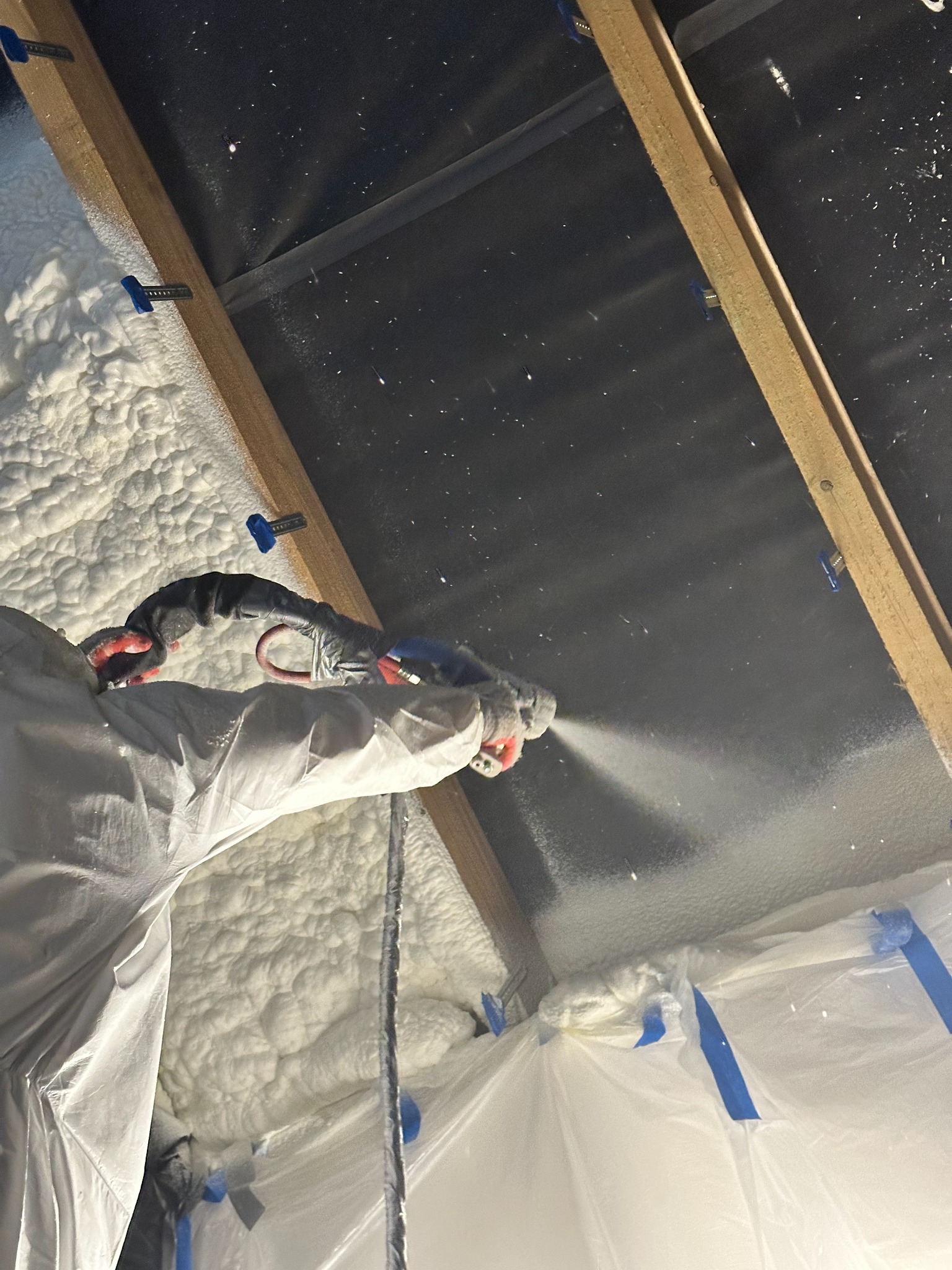 Aplikacja pianki izolacyjnej PUR na poddaszu, widoczne elementy konstrukcji dachu i zabezpieczenia folią. Praca w toku, piana pokrywa powierzchnię.