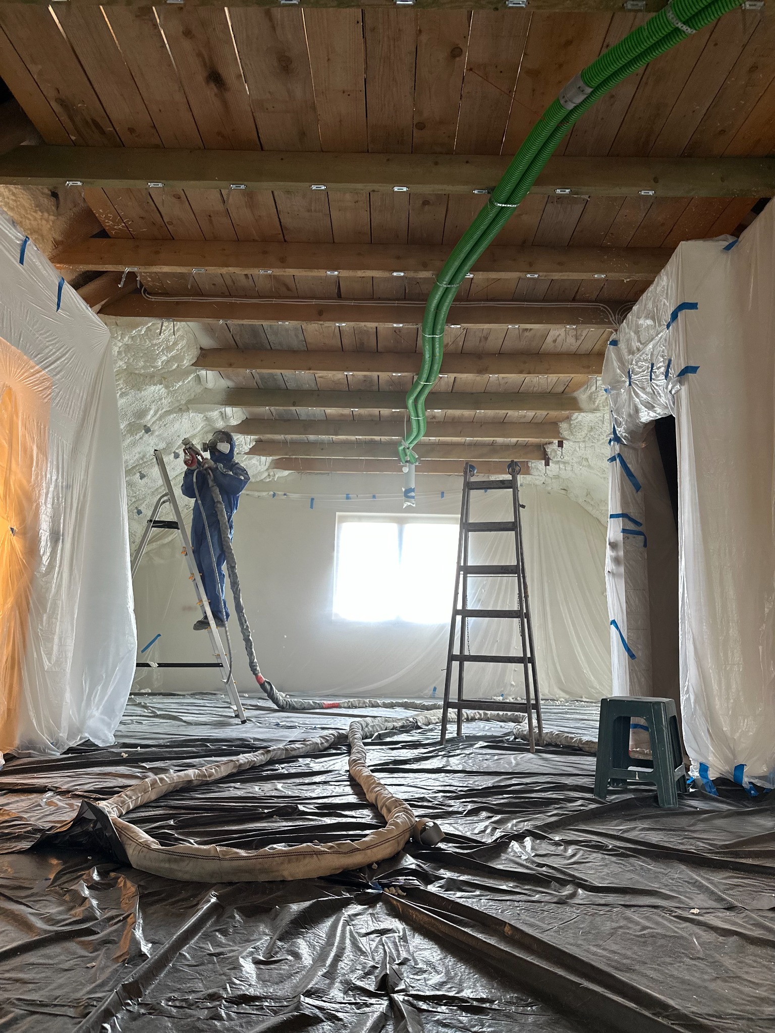 Prace izolacyjne poddasza: aplikacja piany natryskowej przez pracownika w kombinezonie, widoczne zabezpieczenia folią, drewniana konstrukcja dachu i zielone węże doprowadzające materiał.