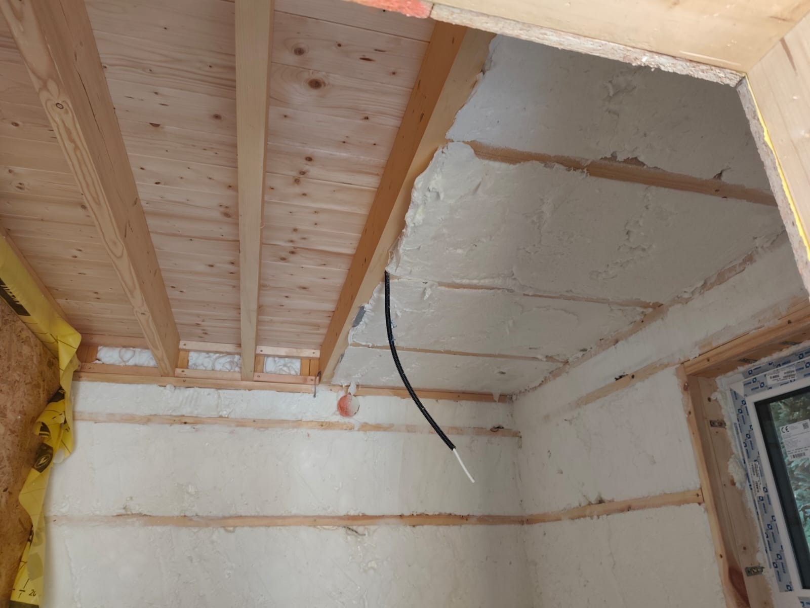 Wnętrze ocieplonego poddasza z widoczną konstrukcją drewnianą i białą warstwą izolacyjną na ścianach. Przewód elektryczny biegnie wzdłuż ocieplonej ściany.