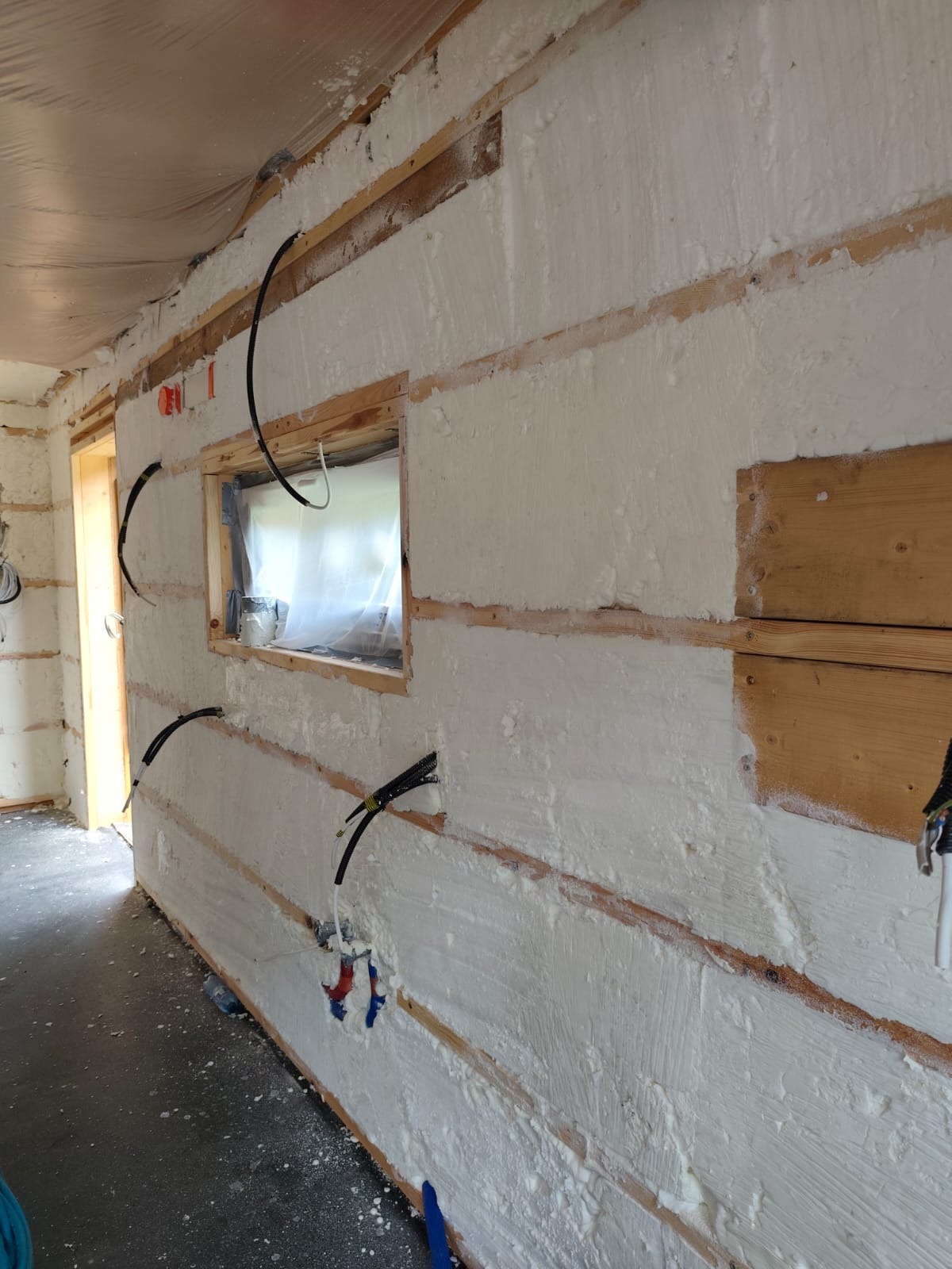 Ocieplony pianą PUR budynek w trakcie prac, widoczne drewniane belki i wystające przewody elektryczne. Okno zabezpieczone folią. Ściana pokryta białą pianą.