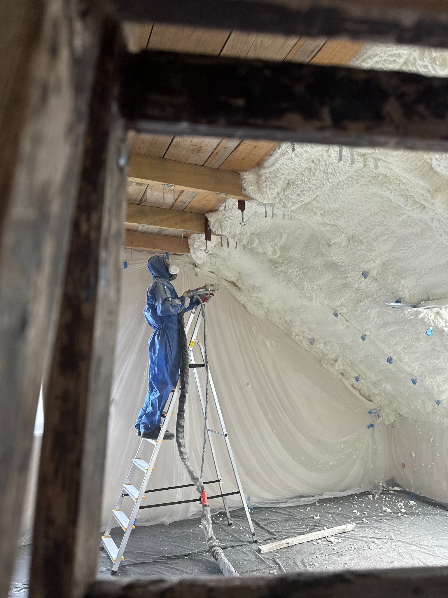 Pracownik w kombinezonie na drabinie aplikuje piankę PUR na poddaszu, widok przez drewnianą ramę. Ocieplenie pianką widoczne na skośnym dachu.