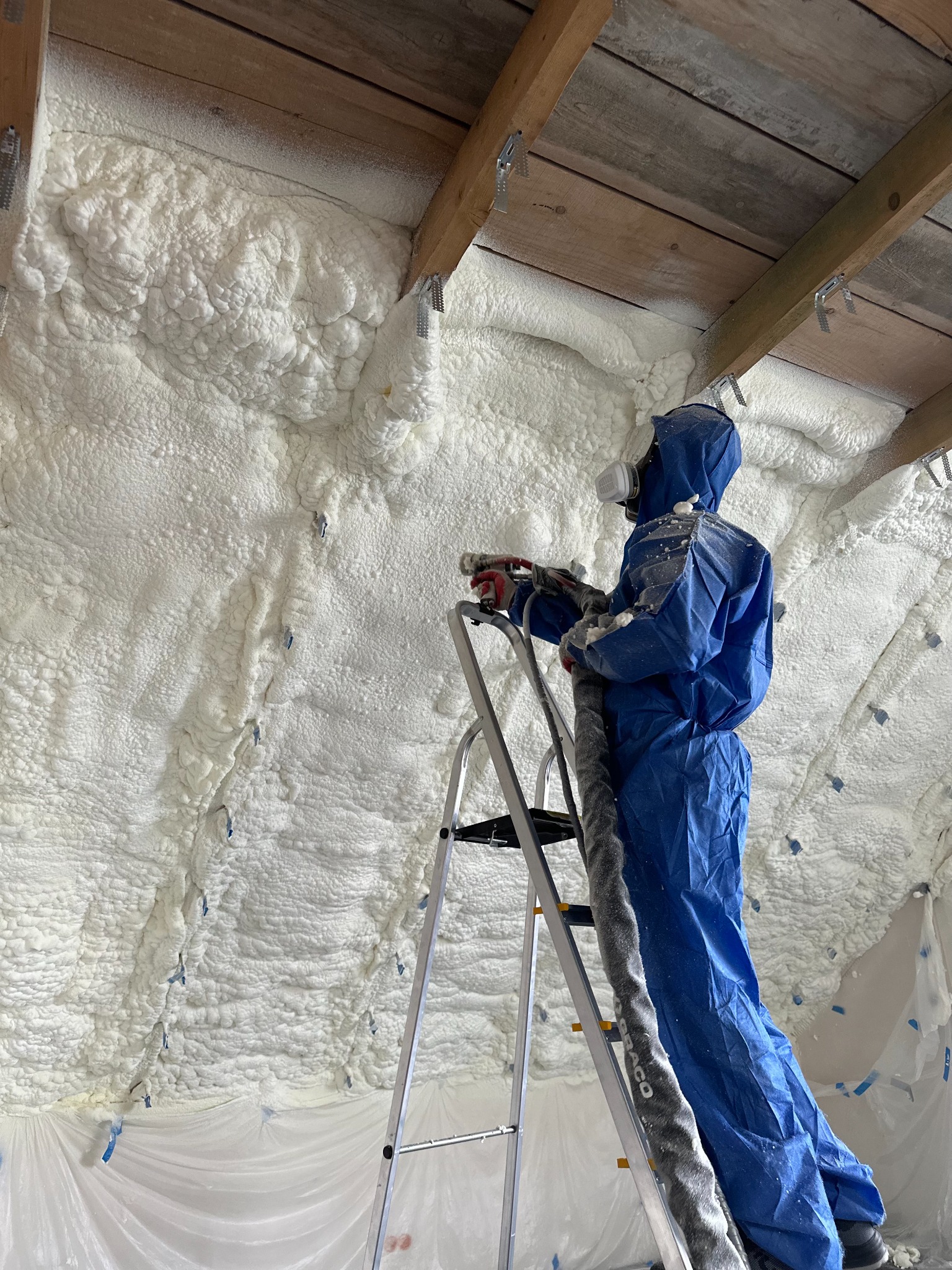 Pracownik w kombinezonie ochronnym aplikuje pianę izolacyjną na poddaszu, stojąc na drabinie. Widoczne drewniane elementy konstrukcyjne dachu i zabezpieczająca folia.