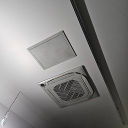 MUSZYN MARCIN MUSZYŃSKI - Sufitowa klimatyzacja kasetonowa z kratką w pomieszczeniu, obok właz rewizyjny, widoczne elementy instalacji elektrycznej.