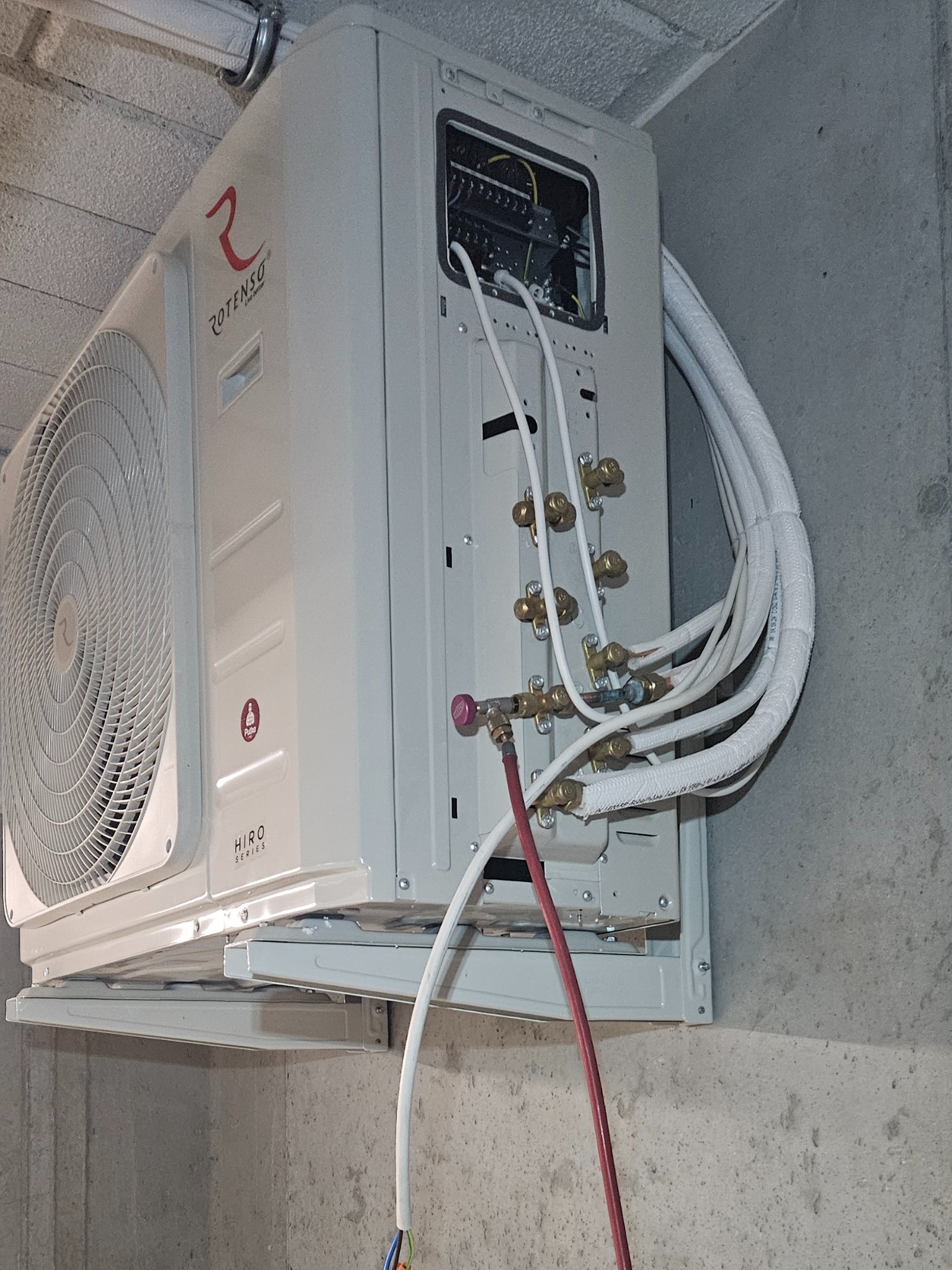 Biała jednostka klimatyzacyjna Rotenso Hiro Series zamontowana na szarej, surowej ścianie, z widocznymi rurami i kablami podłączeniowymi w trakcie instalacji.