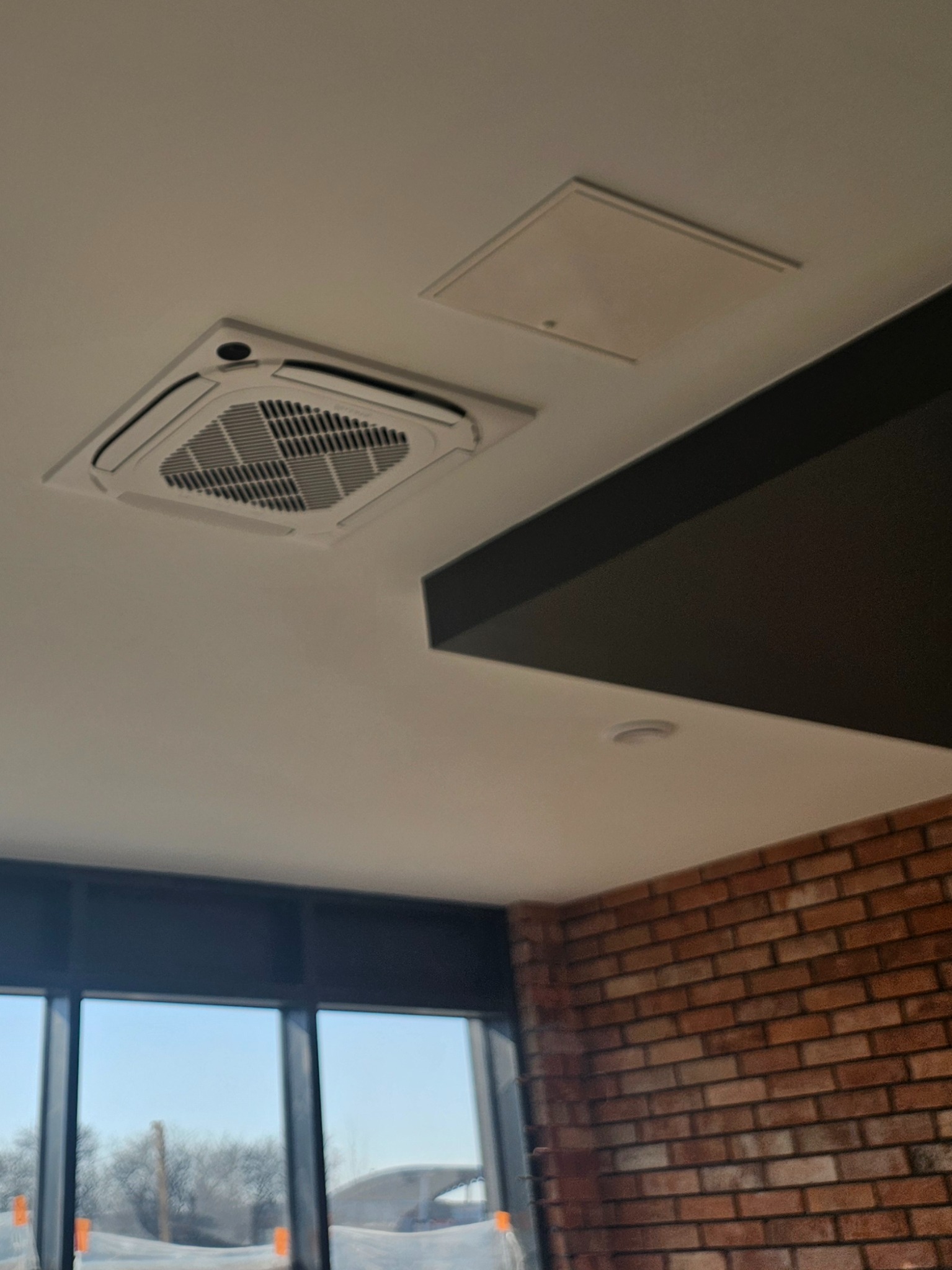 Kasetonowa klimatyzacja sufitowa w biurze z widoczną kratką wentylacyjną i panelem dostępowym, na tle ceglanej ściany i okna z widokiem na zimowy krajobraz.
