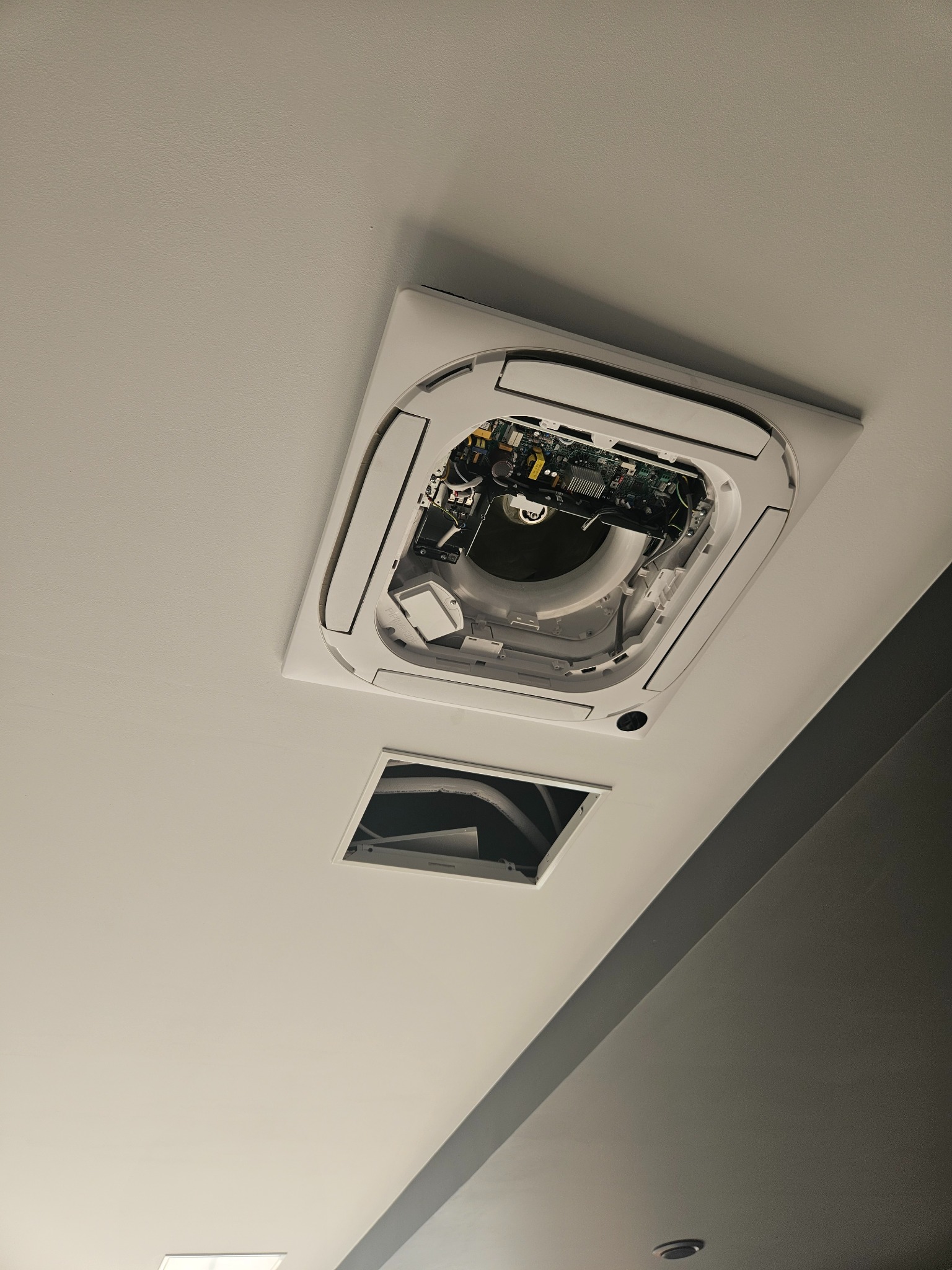 Otwarty sufit podwieszany z widoczną instalacją klimatyzacji kasetonowej, w trakcie serwisu, z widoczną elektroniką i elementami mechanicznymi, dodatkowy otwór w suficie obok.