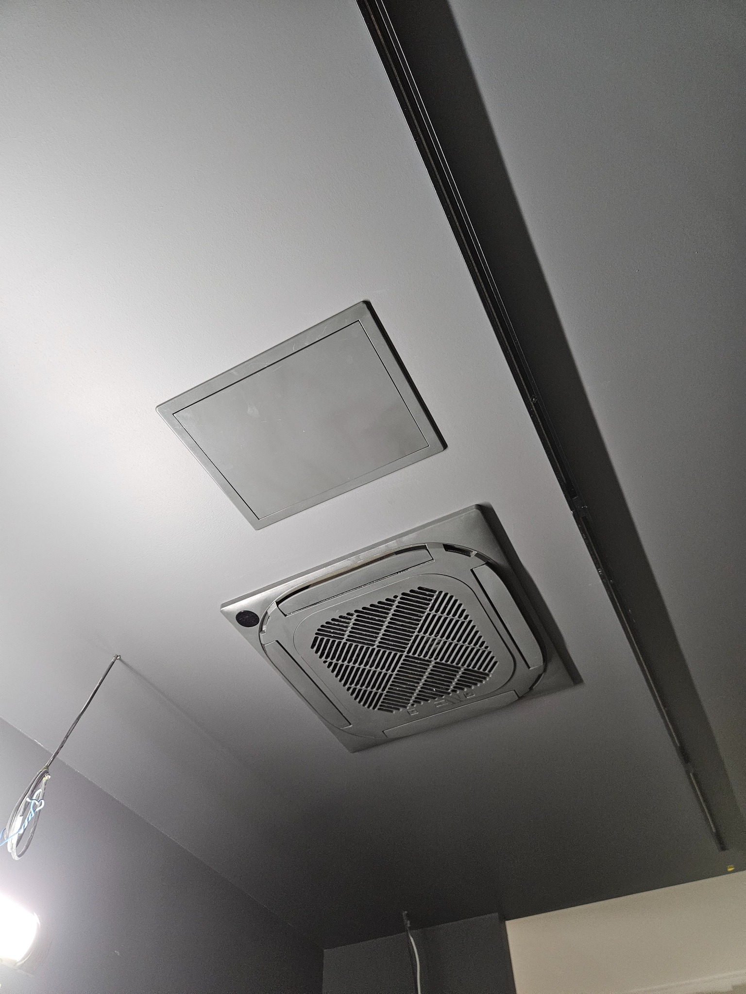 Sufitowa klimatyzacja kasetonowa z kratką w pomieszczeniu, obok właz rewizyjny, widoczne elementy instalacji elektrycznej.