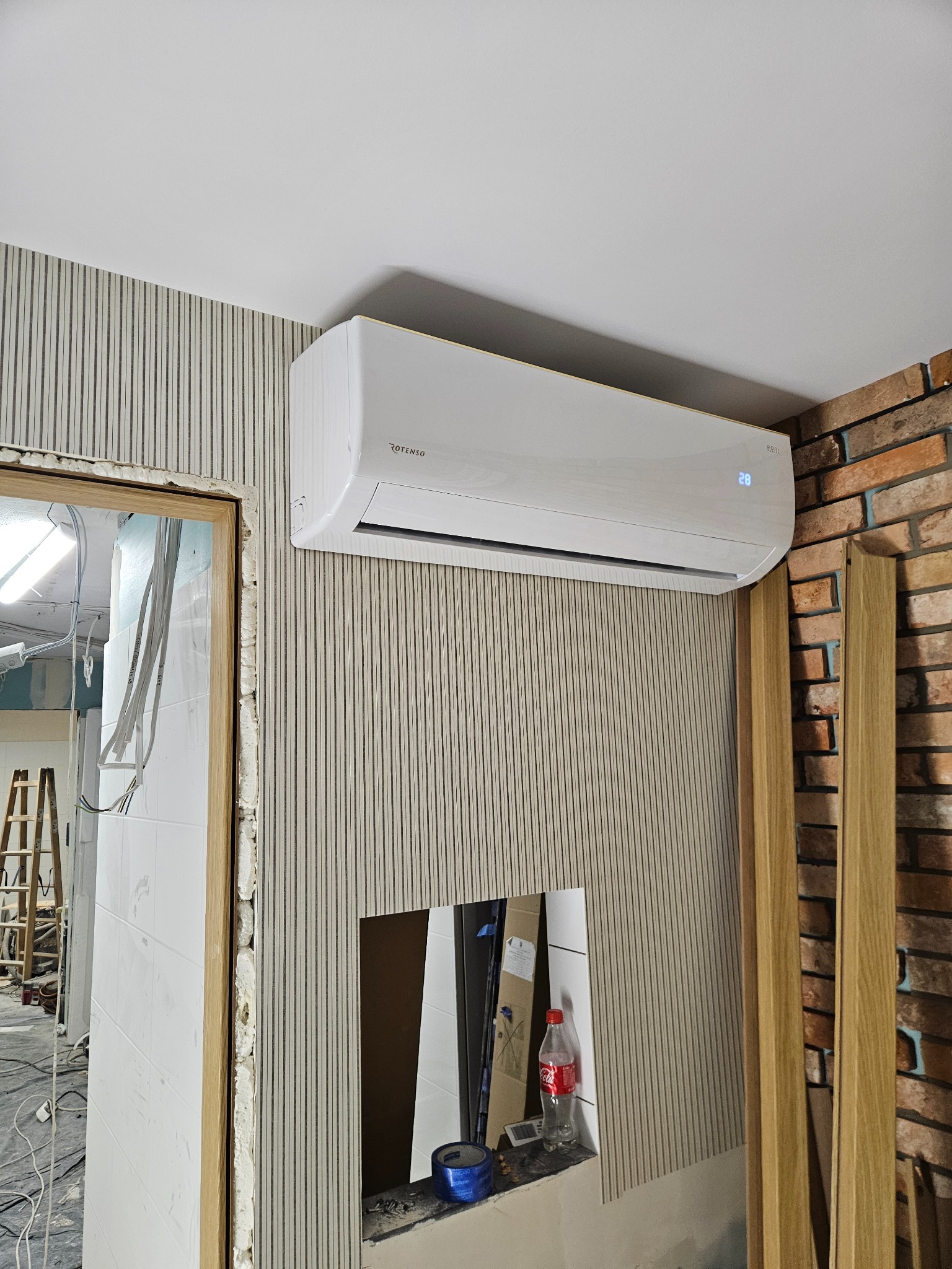 Biały klimatyzator Rotenso zamontowany na ścianie z dekoracyjnymi panelami w pionowe paski, obok fragmentu ściany z cegły i niedokończonego otworu drzwiowego z widoczną pianką montażową.