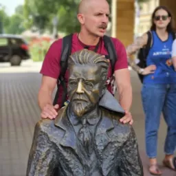 Mężczyzna z wąsami obejmuje ramionami brązową rzeźbę popiersia mężczyzny w okularach i marynarce, w tle kobieta w sportowej koszulce i okulary.