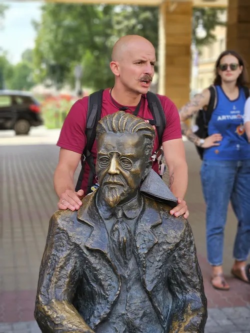 Mężczyzna z wąsami obejmuje ramionami brązową rzeźbę popiersia mężczyzny w okularach i marynarce, w tle kobieta w sportowej koszulce i okulary.