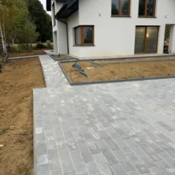 Nowoczesny dom z szarą kostką brukową na podjeździe i ścieżce. Widoczne rabaty ziemne z niebieskimi rurami. Minimalistyczny styl, stonowana kolorystyka.