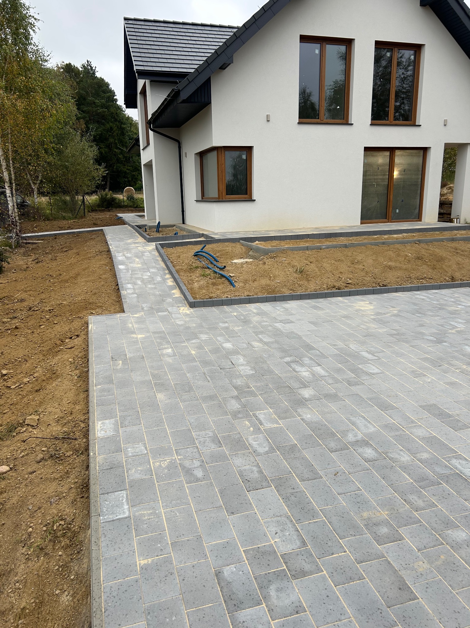 Nowoczesny dom z szarą kostką brukową na podjeździe i ścieżce. Widoczne rabaty ziemne z niebieskimi rurami. Minimalistyczny styl, stonowana kolorystyka.