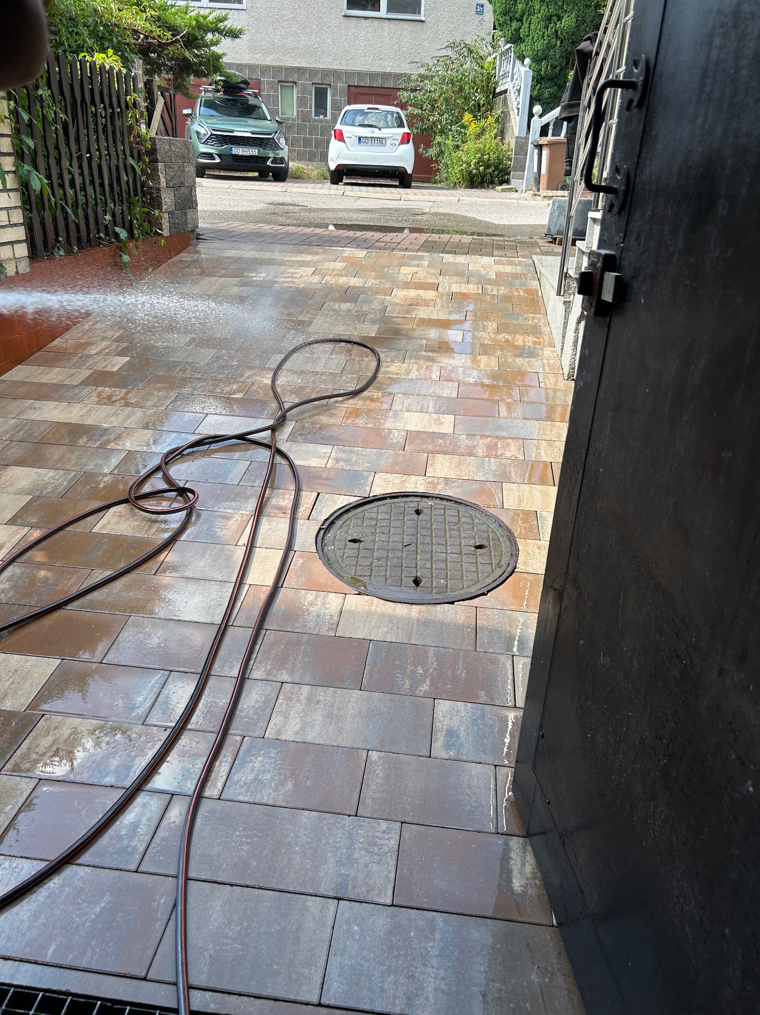 Czyszczenie kostki brukowej wodą pod ciśnieniem na podjeździe. Wąż ogrodowy leży na mokrej powierzchni. W tle dwa samochody i budynek.