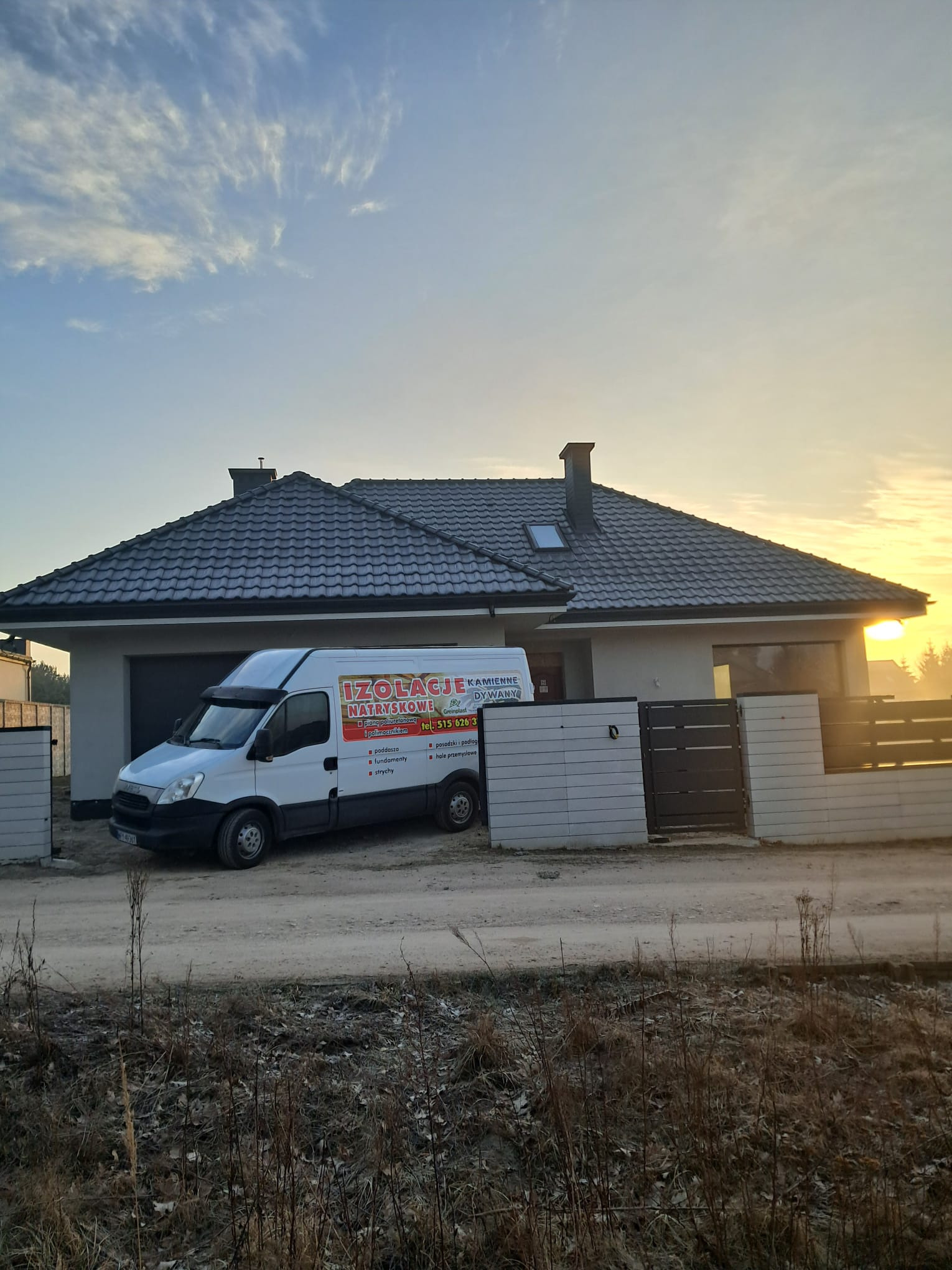 Biały samochód dostawczy z reklamą firmy izolacyjnej zaparkowany przed nowoczesnym domem jednorodzinnym z ciemną dachówką, w tle wschodzące słońce.