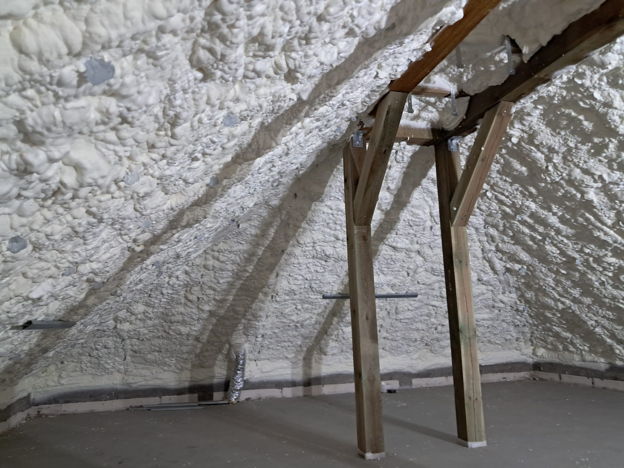 Poddasze z drewnianą konstrukcją pokryte grubą warstwą białej pianki izolacyjnej, widoczne fragmenty ściany z szarymi bloczkami i element wentylacji.