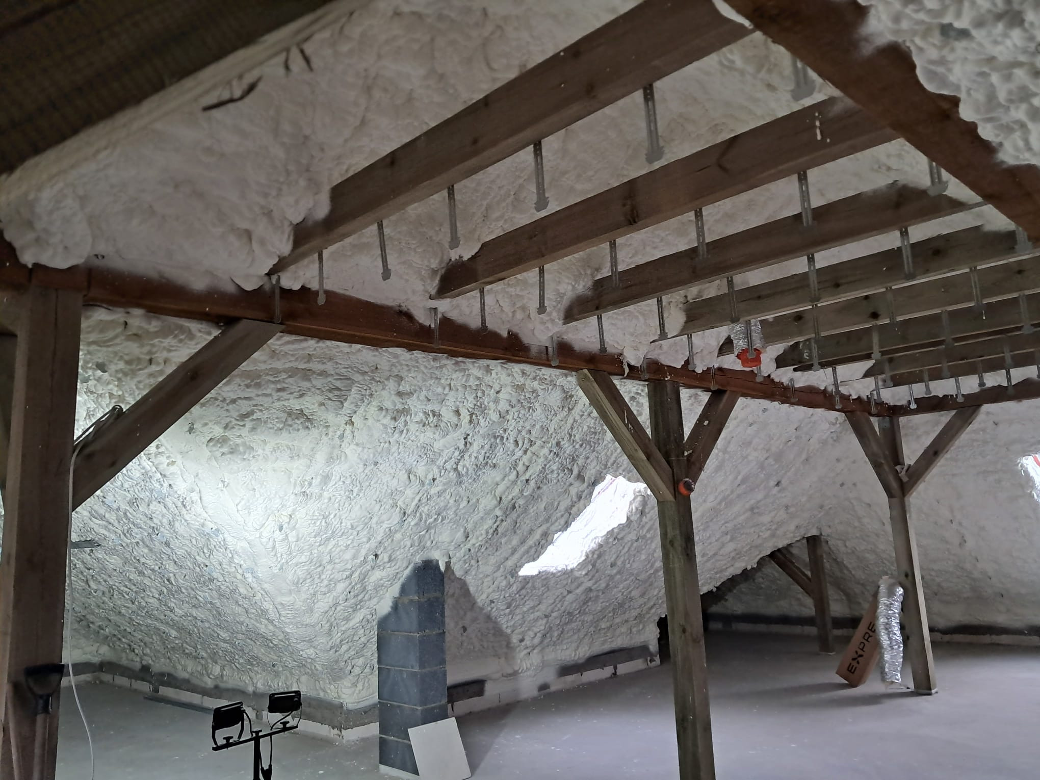 Poddasze z drewnianą konstrukcją pokryte grubą warstwą białej pianki izolacyjnej, widoczne metalowe elementy montażowe i fragment komina.