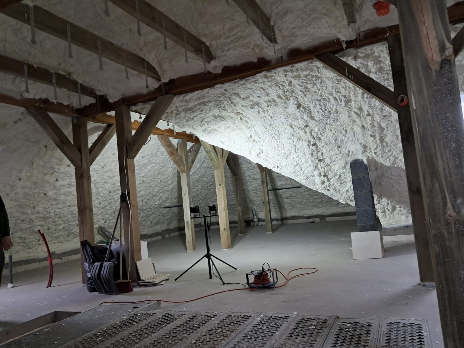Poddasze z drewnianą konstrukcją pokryte białą pianką izolacyjną, widoczne narzędzia pracy i oświetlenie budowlane na statywie.