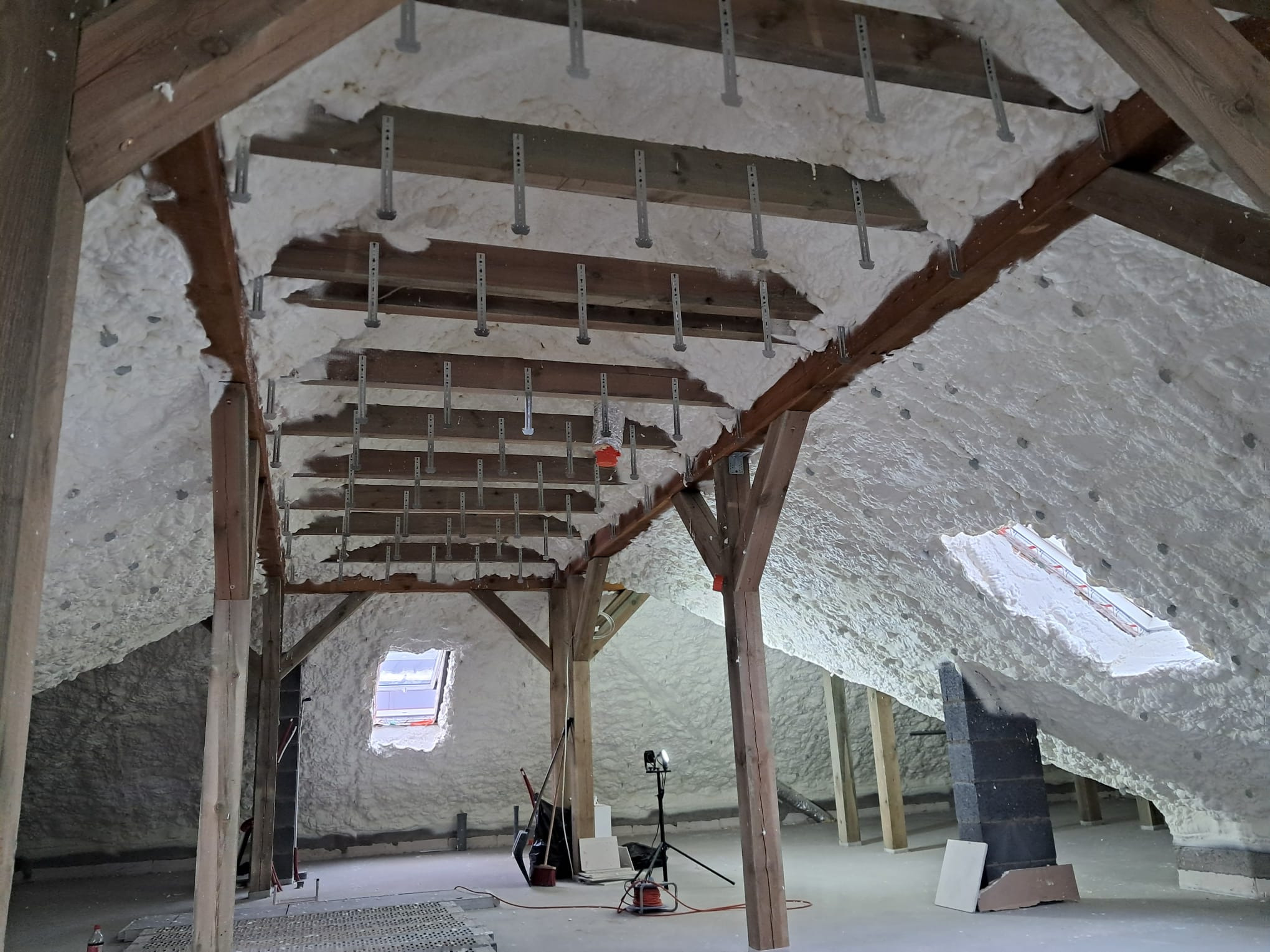 Poddasze ocieplone białą pianką z widocznymi drewnianymi belkami konstrukcyjnymi i metalowymi elementami mocującymi, okno dachowe w tle, narzędzia pracy widoczne na podłodze.