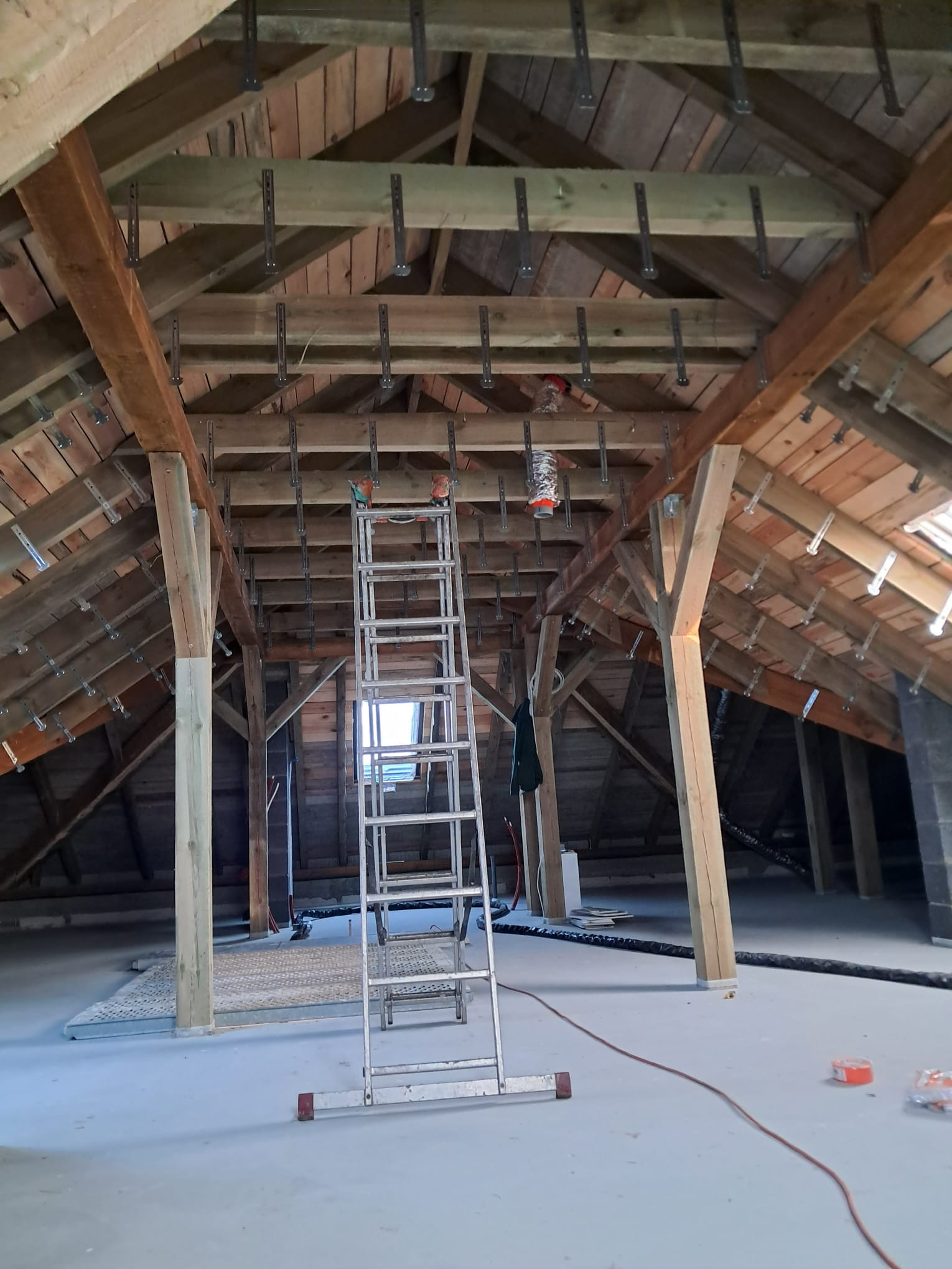 Poddasze w trakcie ocieplania, widoczne drewniane krokwie z zamontowanymi metalowymi uchwytami, aluminiowa drabina na środku pomieszczenia, przygotowane materiały izolacyjne i przewody na podłodze.