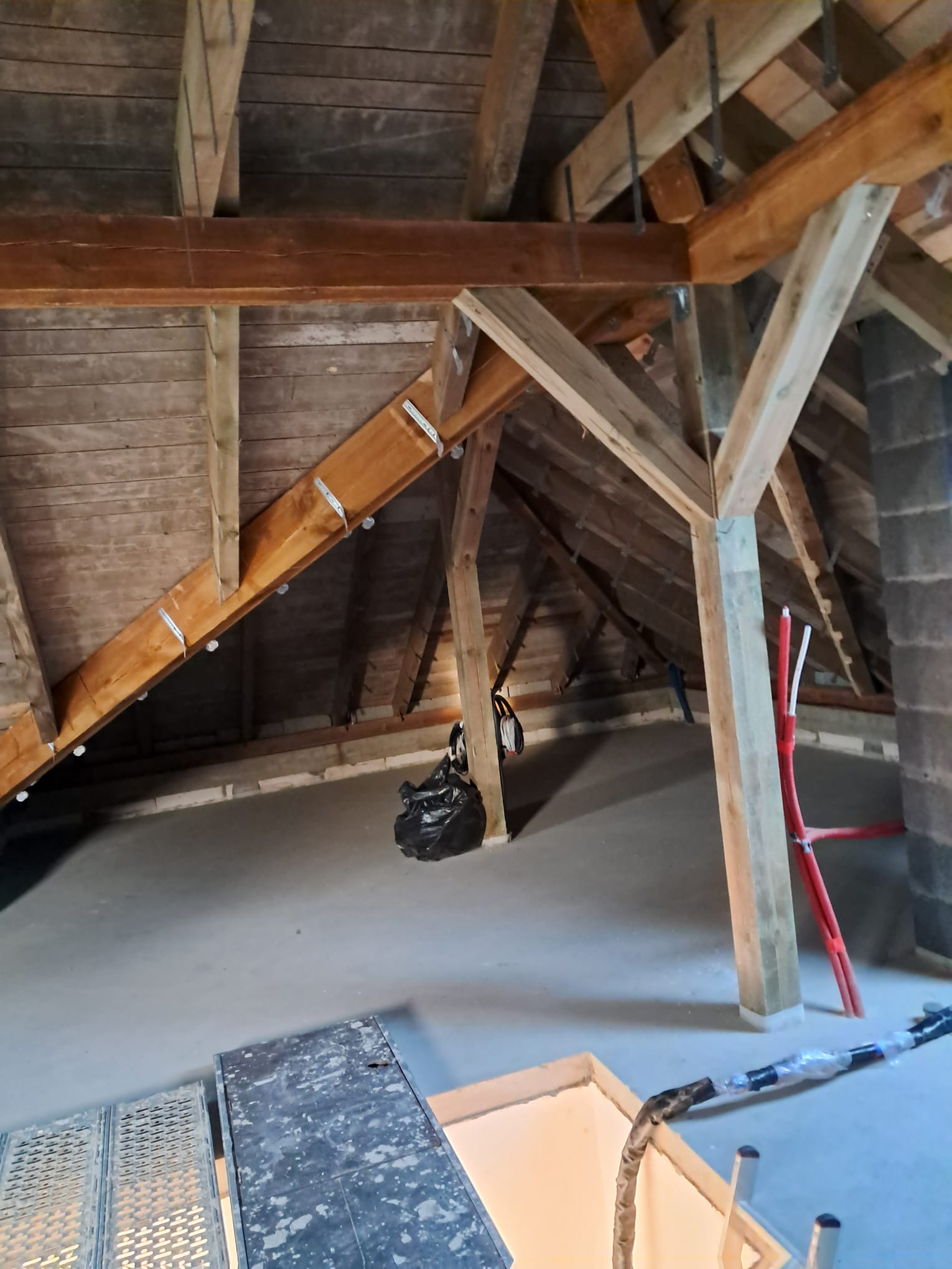 Poddasze z drewnianą konstrukcją więźby dachowej, widoczne krokwie i słupy, przygotowane do prac izolacyjnych, czarny worek na śmieci i czerwone rury w tle.
