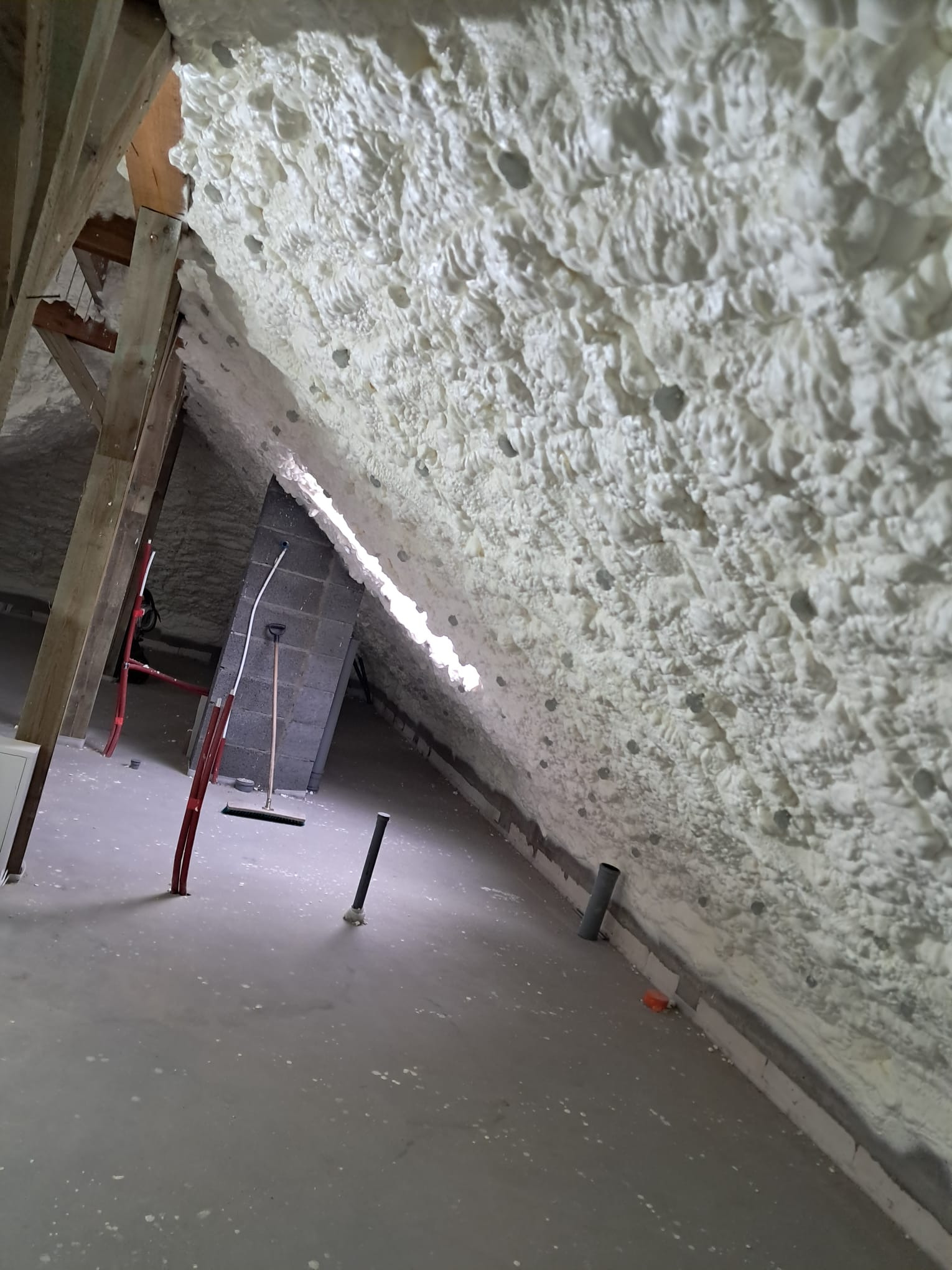 Poddasze z widoczną warstwą białej, natryskowej izolacji termicznej, drewnianą konstrukcją więźby dachowej, kominem z cegły i rozstawioną drabiną.