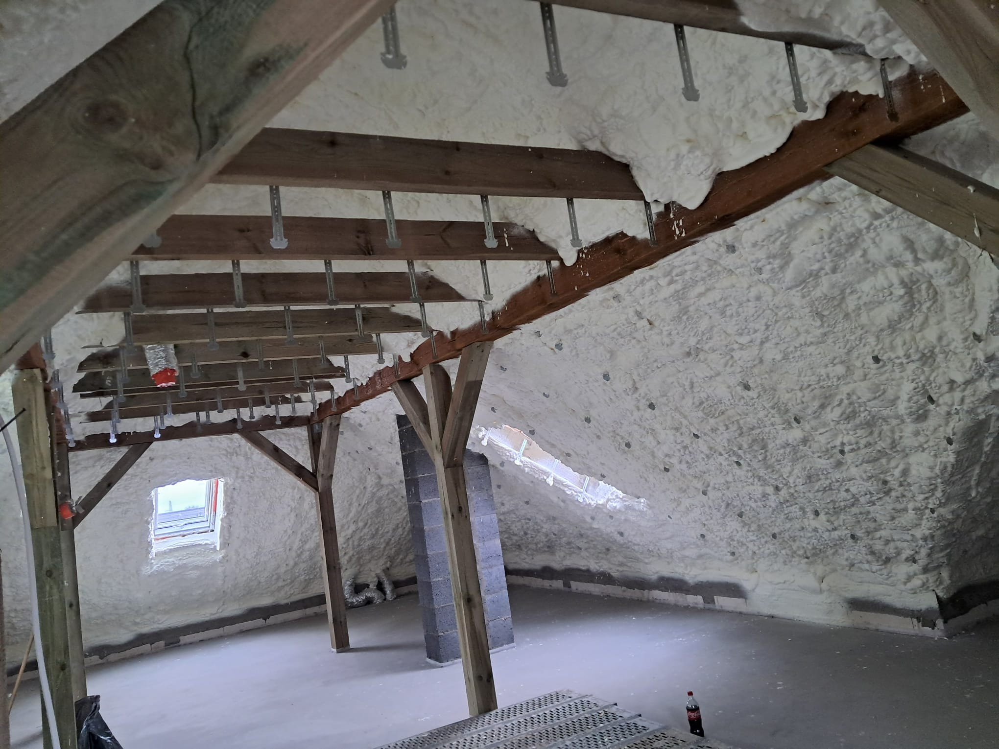 Poddasze po natrysku pianki izolacyjnej, widoczne drewniane elementy konstrukcyjne i wystające metalowe mocowania, fragment okna dachowego i butelka Coca-Coli w rogu.