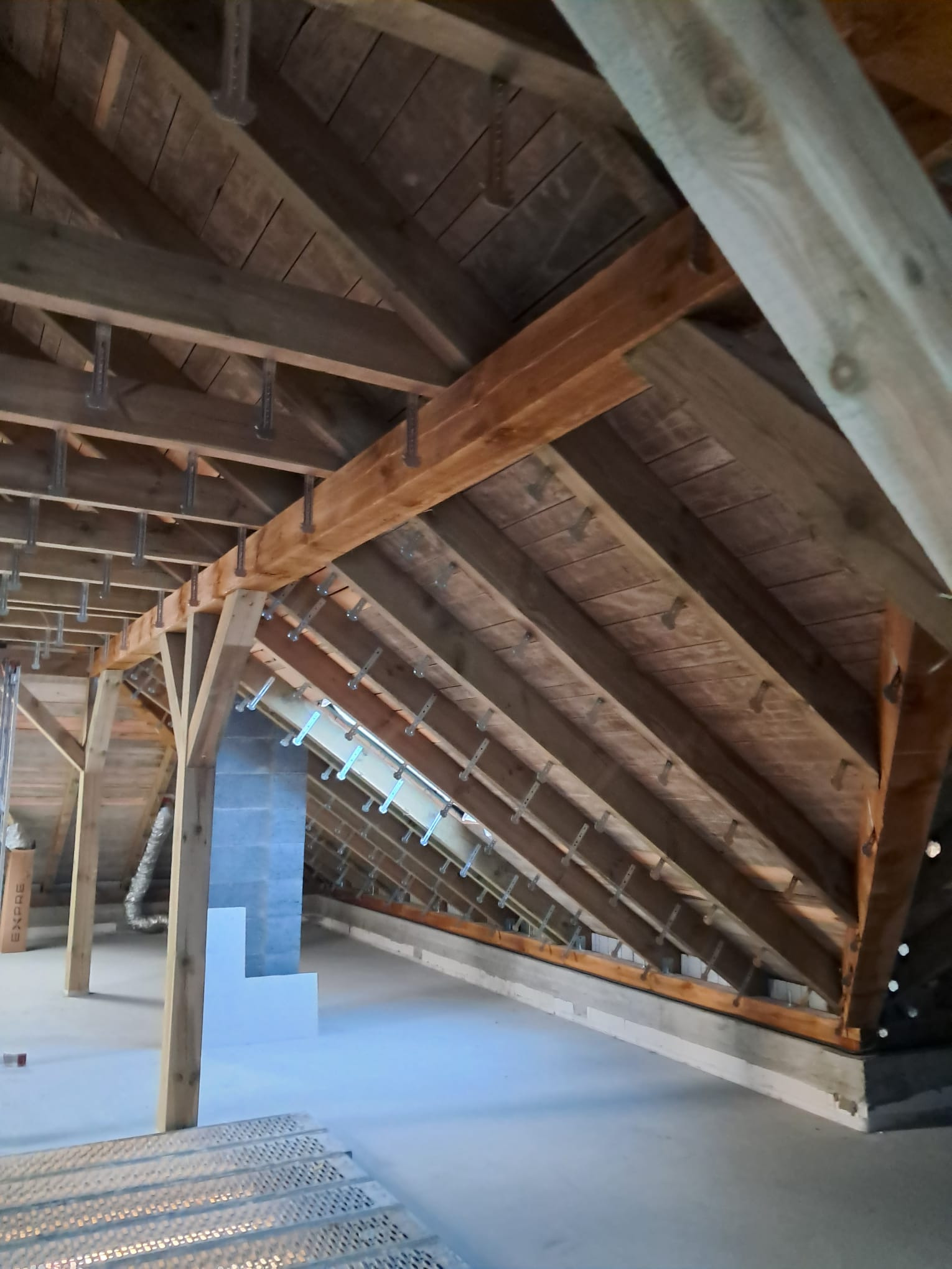 Drewniana konstrukcja dachu poddasza z widocznymi krokwiami, łatami i elementami mocującymi, przygotowana do prac izolacyjnych, z fragmentem rusztowania na pierwszym planie i kominem w tle.