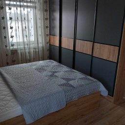 KONRAD-MEBLE - Sypialnia z łóżkiem i przesuwną szafą wnękową z czarnymi drzwiami i drewnianymi panelami, widok na okno z firankami w tle.