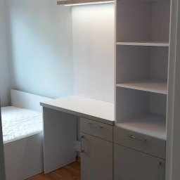 KONRAD-MEBLE - Pokój z zabudową meblową: biurko z szufladami, półki i szafki górne w odcieniach bieli i szarości, dopasowane do łóżka, na podłodze panele.