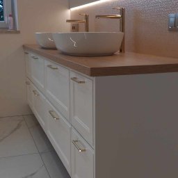 KONRAD-MEBLE - Nowoczesna łazienka z podwójną umywalką nablatową, białe szafki z frezowanymi frontami i złotymi uchwytami, drewniany blat, oświetlenie LED nad lustrem i marmurowe płytki podłogowe.