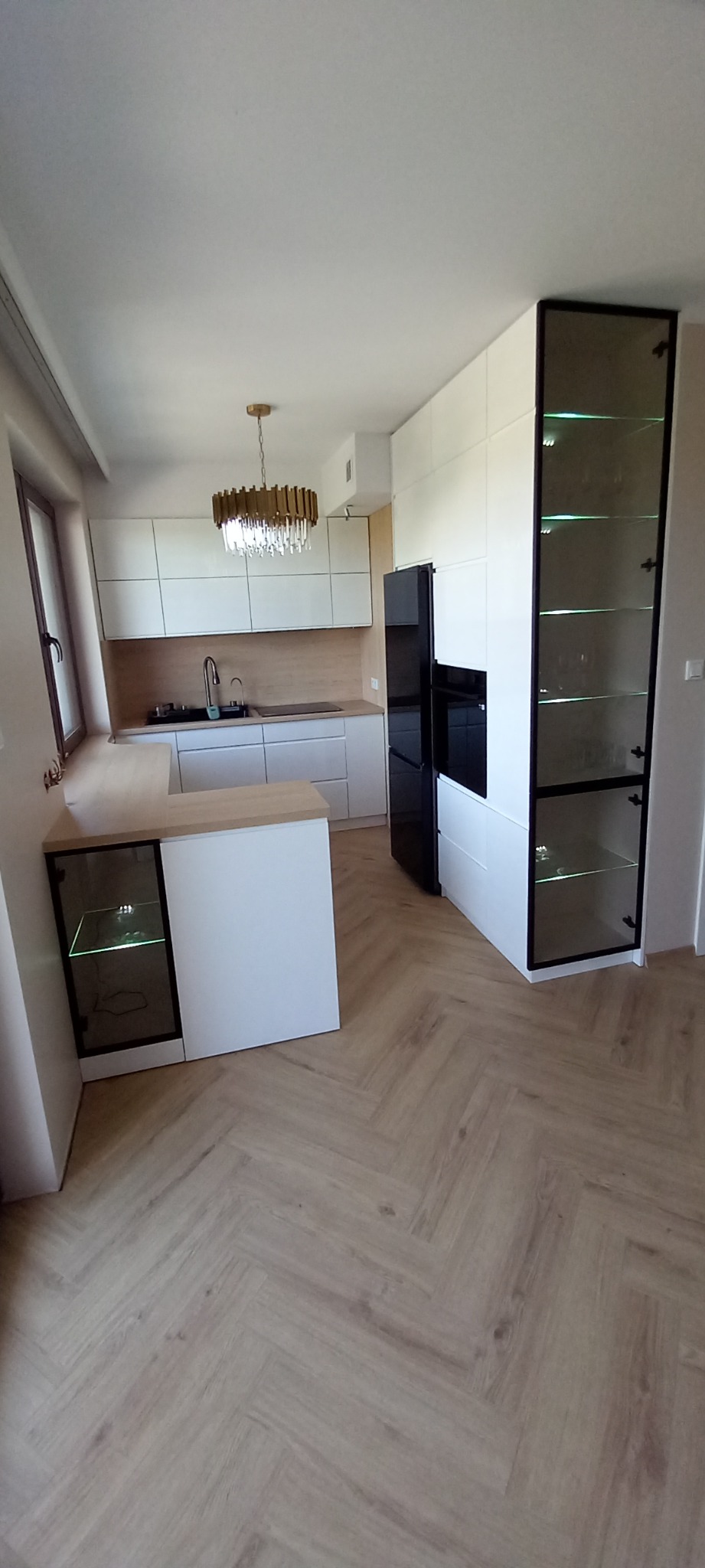 Nowoczesna kuchnia z białymi frontami, drewnianym blatem i podłogą w jodełkę; widoczna wyspa z podświetlaną witryną, czarna lodówka i przeszklona szafka z podświetleniem LED.