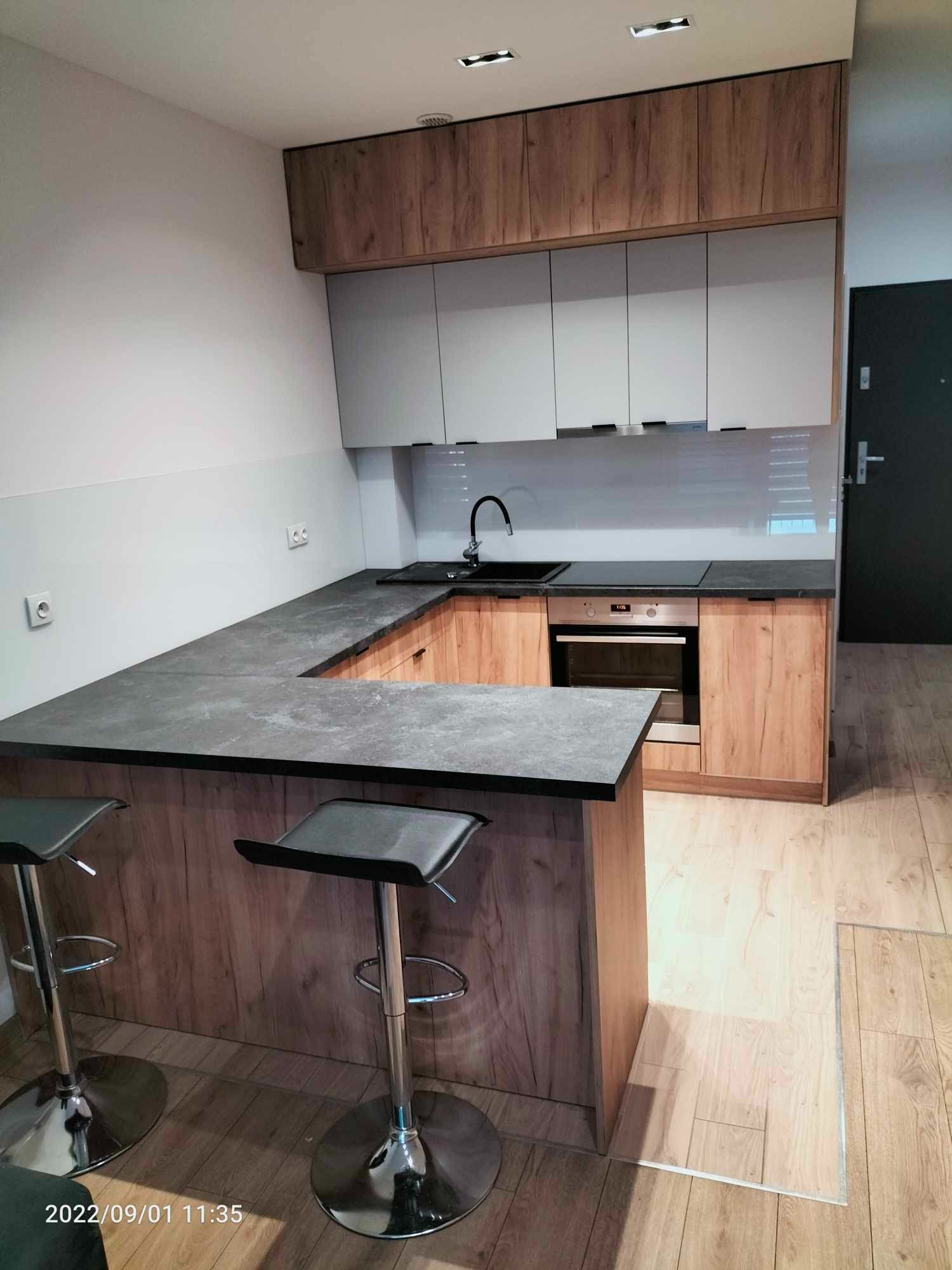 Nowoczesna kuchnia z wyspą, szare szafki wiszące i drewnopodobne szafki dolne, czarny blat i dwa chromowane hokery na drewnianej podłodze.