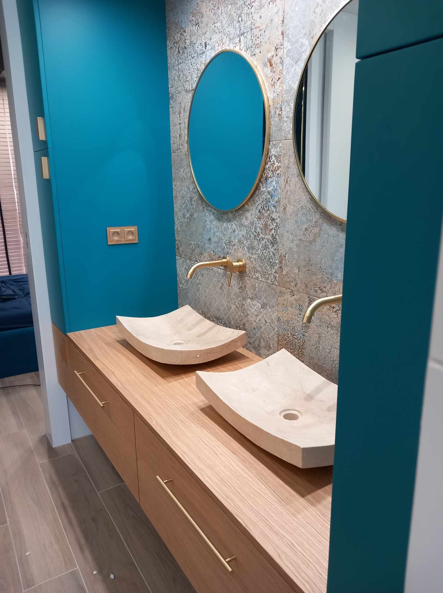 Nowoczesna łazienka z dwiema kamiennymi umywalkami w kształcie muszli, złotą armaturą i okrągłymi lustrami na tle wzorzystych płytek, szafka z drewnianymi frontami i długimi uchwytami, obok...