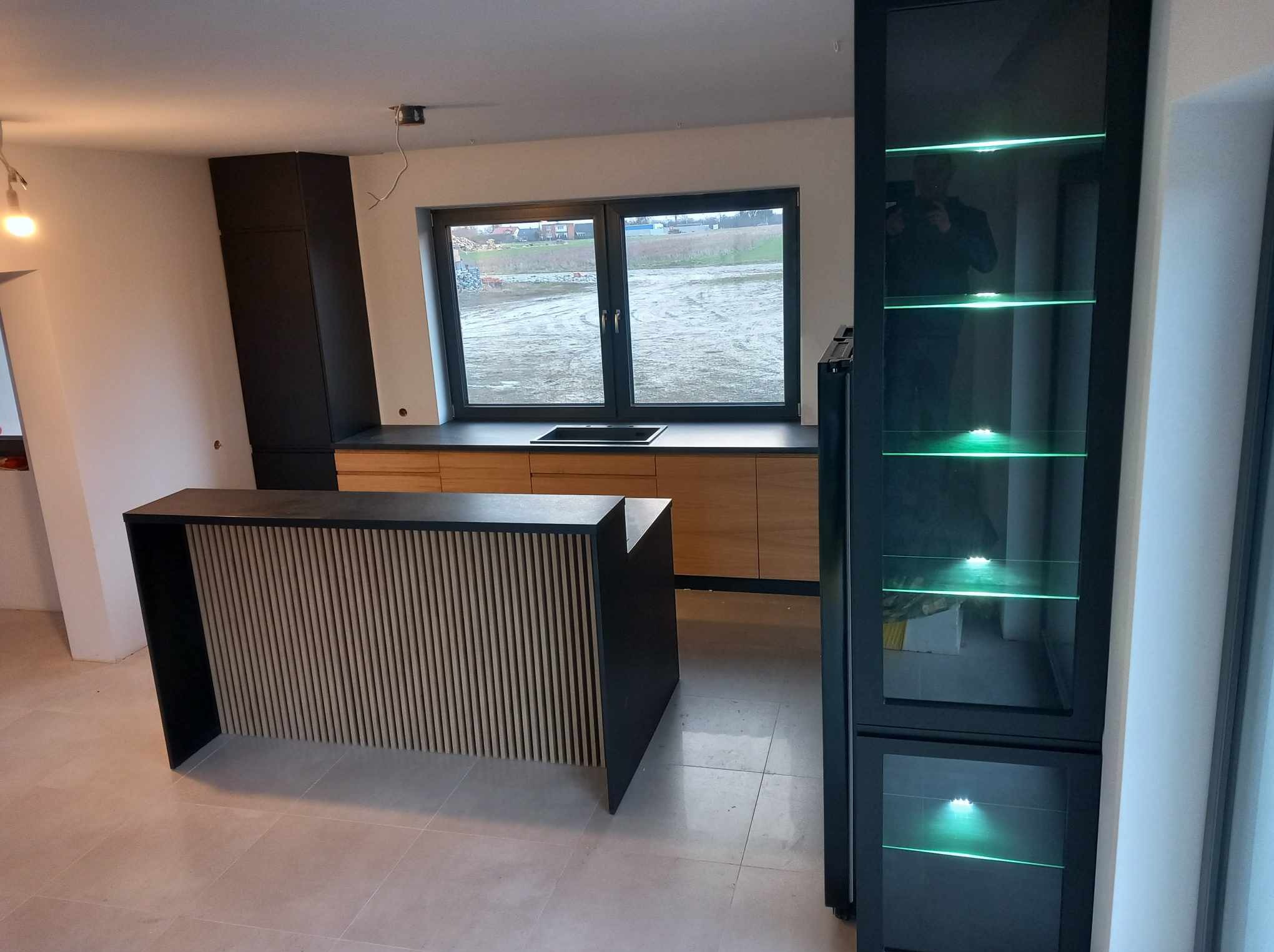 Nowoczesna kuchnia z drewnianymi szafkami, czarnym blatem i wyspą z lamelami, oświetlona szafka z przeszklonym frontem, widok na pole z okna.